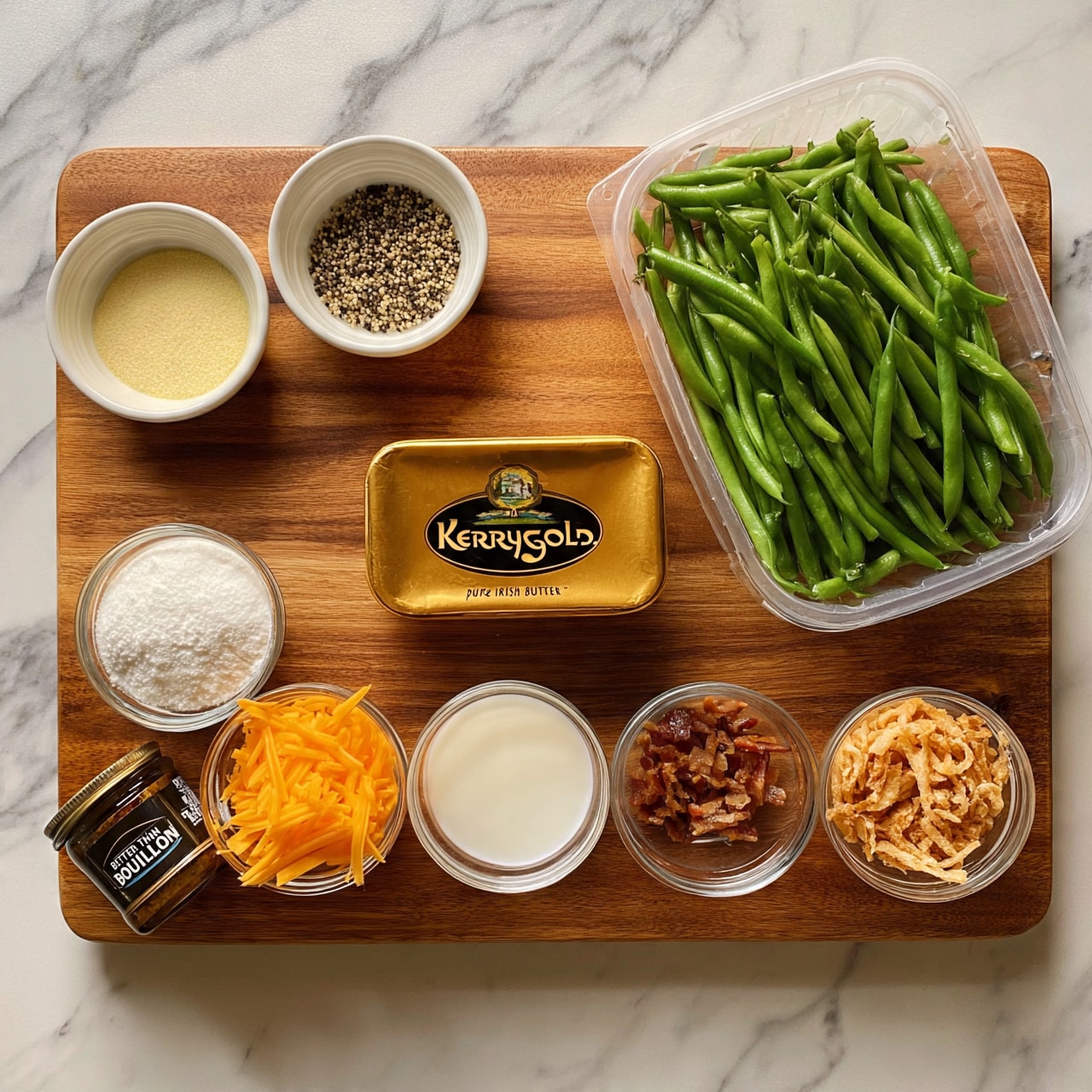 square image The image shows nine ingredients arranged neatly on a wooden cutting board placed on a white marbled texture. At the top right, there is a clear plastic container filled with bright green fresh green beans. To the left of it, two small white bowls hold a pale yellow granular ingredient and a mix of black and white pepper. Below these bowls is a rectangular gold-colored container of Kerrygold Pure Irish Butter. At the bottom left of the butter are four small round clear bowls. From left to right, they contain white powder, shredded orange cheddar cheese, white liquid (likely milk or cream), small pieces of cooked bacon, and light brown fried onions. Next to the bowl of cheese is a small dark glass jar labeled