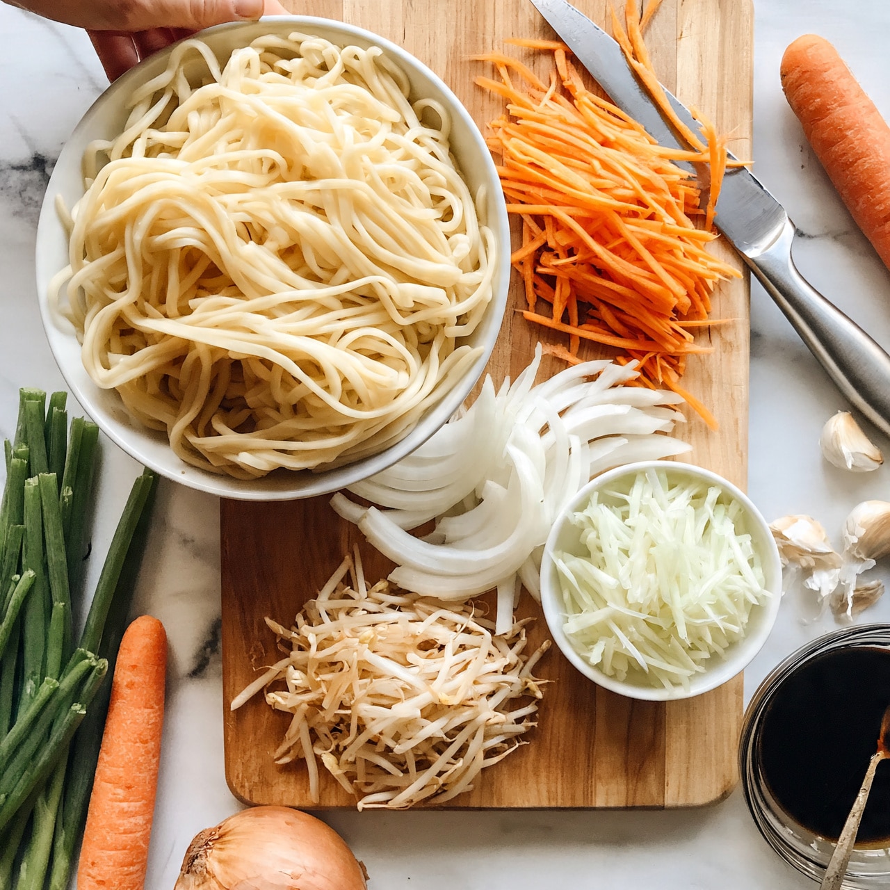 square image The image shows a top-down view of a wooden board with several layers of colorful ingredients arranged neatly. On the left, there is a large white bowl filled with thick, pale yellow noodles. To the right of the noodles, thin orange carrot strips lie in a pile, with some scattered near a silver knife placed diagonally. Below the carrots, white onion slices fan out, and a small white bowl filled with pale green shredded vegetables sits next to them. At the bottom of the board, there is a cluster of light beige bean sprouts. Surrounding the board on a white marbled surface are several green scallions on the left, a whole orange carrot, a brown onion, garlic cloves, and a glass bowl filled with dark soy sauce with a spoon resting inside. In the upper left corner, a woman's hand is seen holding the edge of the board. photo taken with an iphone --ar 4:5 --v 7