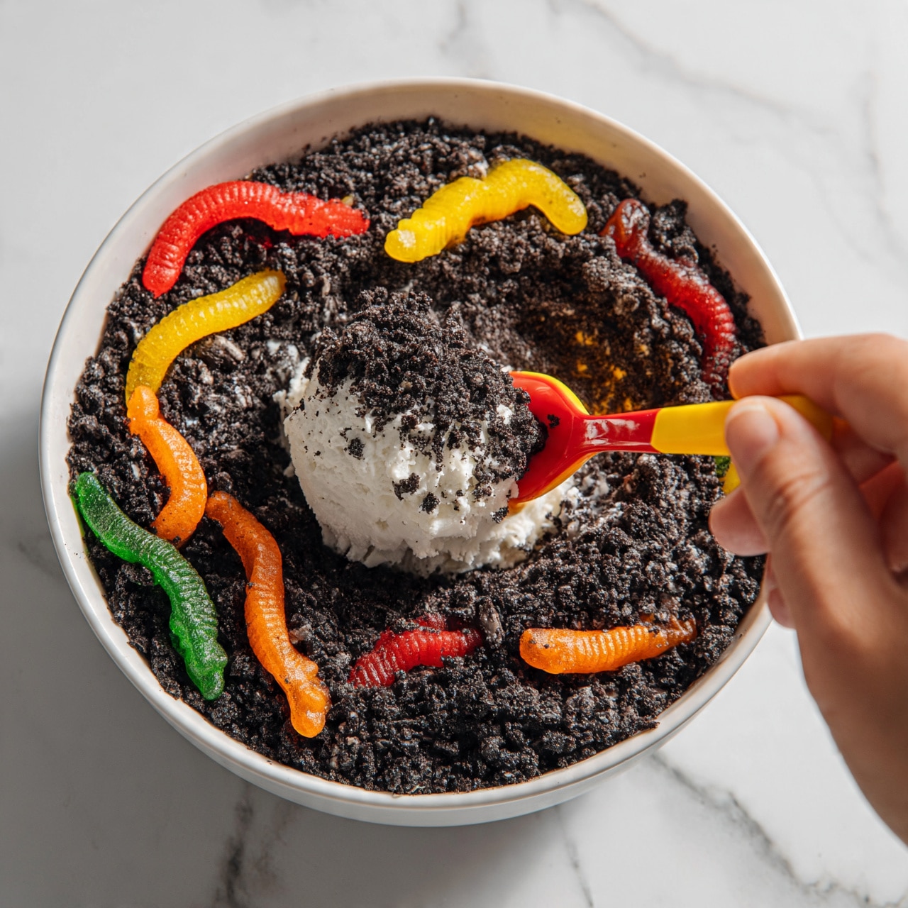 square image A white bowl filled with crushed dark chocolate cookie crumbs forming the bottom layer, scattered with colorful gummy worms in red, yellow, orange, and green on top. In the middle, a scoop of white creamy filling is covered with more dark cookie crumbs, held by a yellow and red spoon being lifted by a woman's hand. The scene is set on a white marbled surface. photo taken with an iphone --ar 4:5 --v 7
