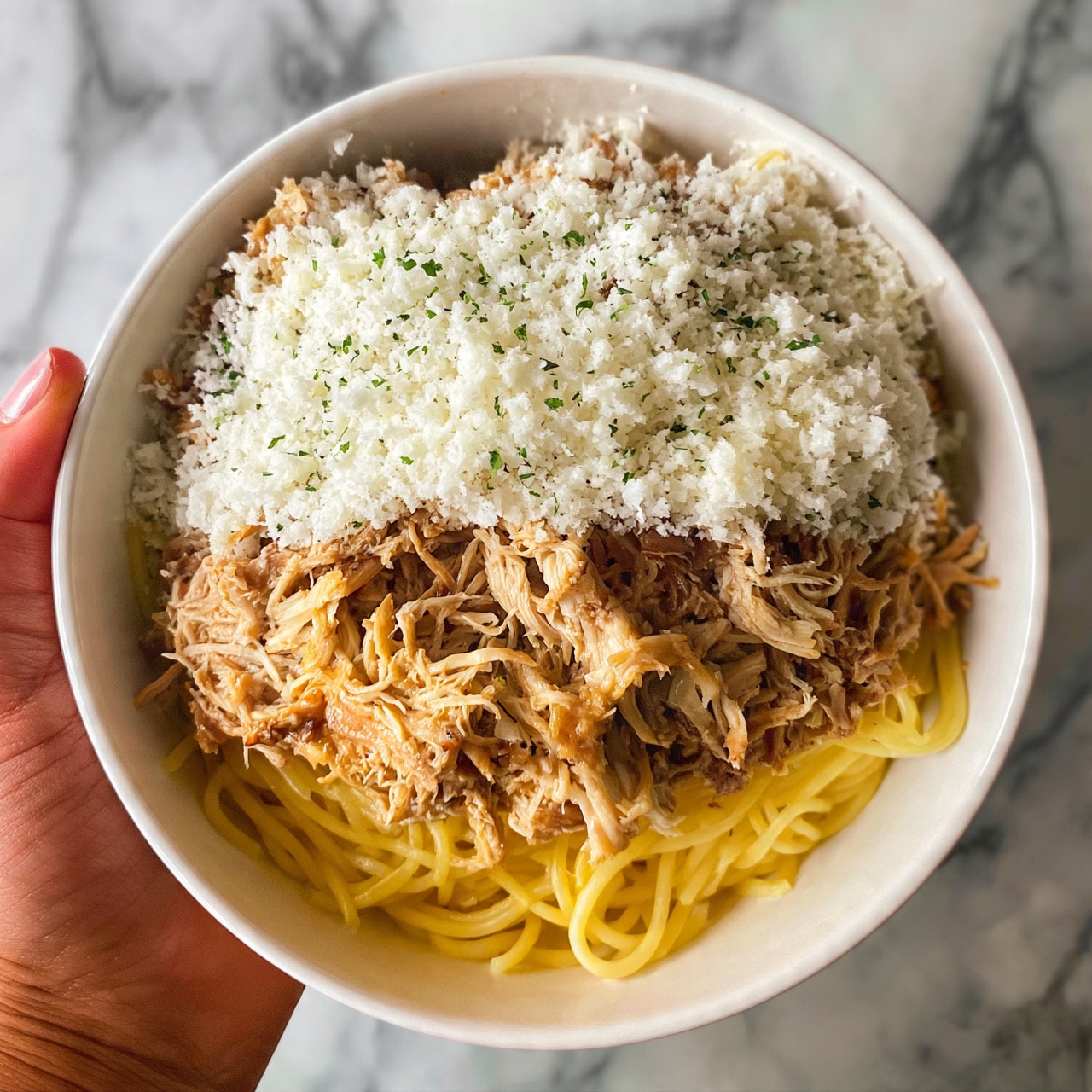 square image A white bowl filled with yellow spaghetti noodles at the bottom, topped with a generous layer of shredded light brown chicken. Above the chicken, there is a layer of white grated cheese sprinkled with small green herb bits. A woman's hand is holding the bowl from the side, and the background shows a white marbled texture. photo taken with an iphone --ar 4:5 --v 7