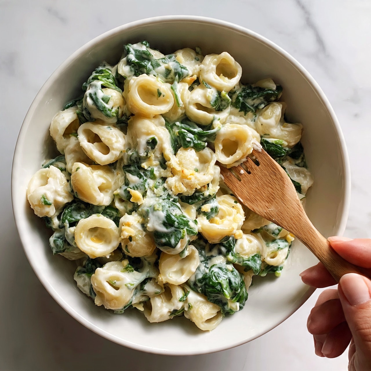 square image A white bowl filled with creamy pasta mixed with green spinach leaves. The pasta is coated in a thick, white sauce with bits of cheese scattered on top. A wooden fork rests inside the bowl, with a woman's hand holding it. The bowl sits on a white marbled surface. photo taken with an iphone --ar 4:5 --v 7