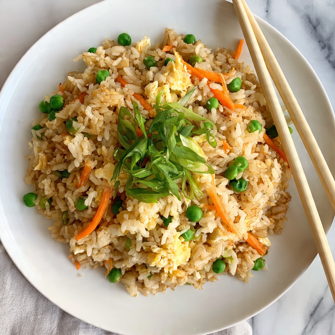 square image A white plate holds a mound of fried rice with visible layers of light brown cooked rice mixed with small bright green peas and thin orange carrot strips. There are bits of soft yellow scrambled egg mixed throughout. On top, a sprinkle of thinly sliced green onions adds a fresh green layer. To the right of the plate, a pair of light-colored wooden chopsticks rests diagonally. The background is a white marbled surface with a light gray cloth underneath the plate. Photo taken with an iphone --ar 4:5 --v 7