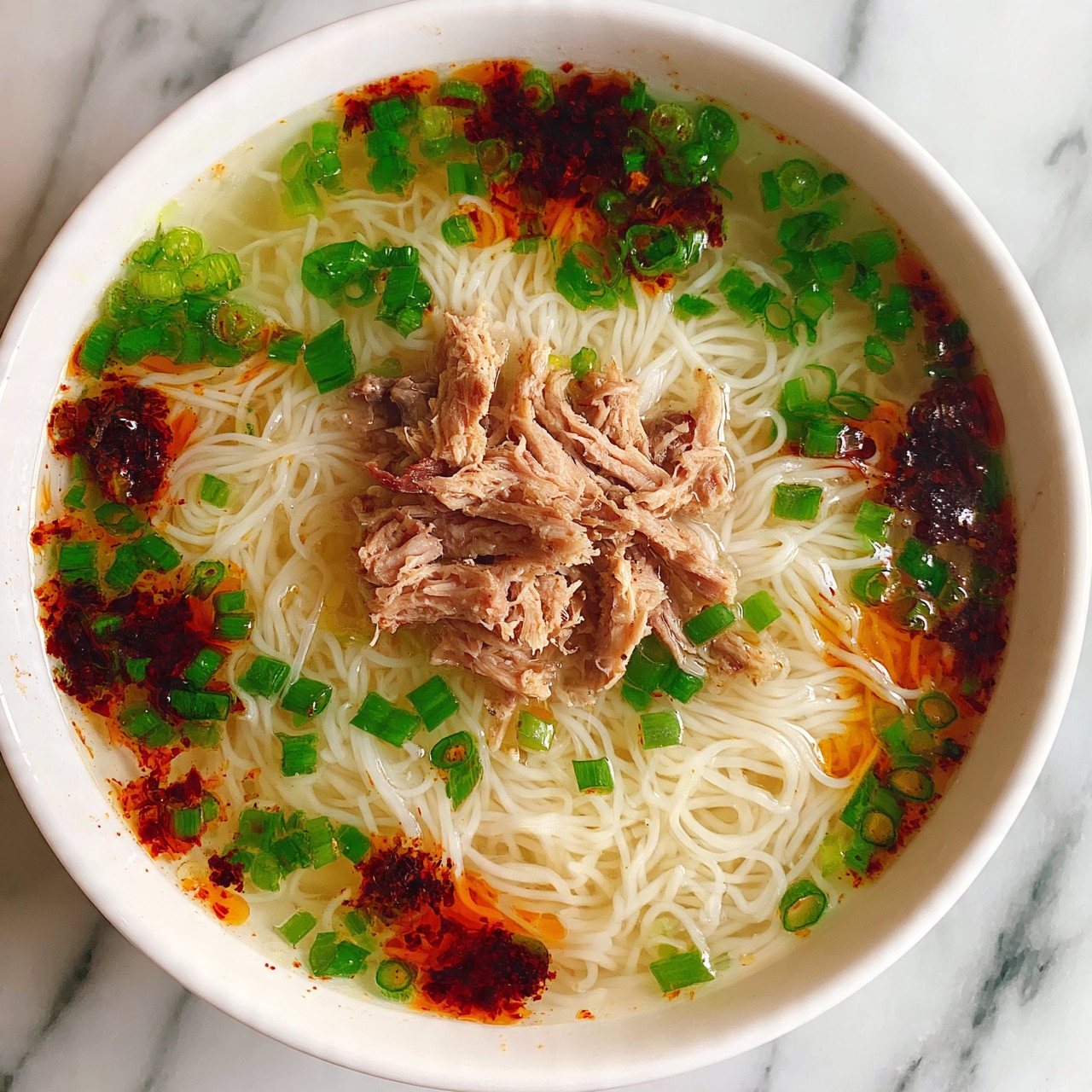 square image The image shows a close-up of a bowl of noodle soup with three main layers. The bottom layer is thin white noodles that fill the bowl. On top of the noodles, there are light brown pieces of shredded meat positioned in the center. Surrounding the meat are bright green chopped scallions scattered evenly across the surface. There are also dark red chili oil spots spread around the bowl, giving a spicy look. The soup's broth looks clear and light, with some oily red patches floating on top. The bowl is white and placed on a white marbled surface. Photo taken with an iphone --ar 4:5 --v 7