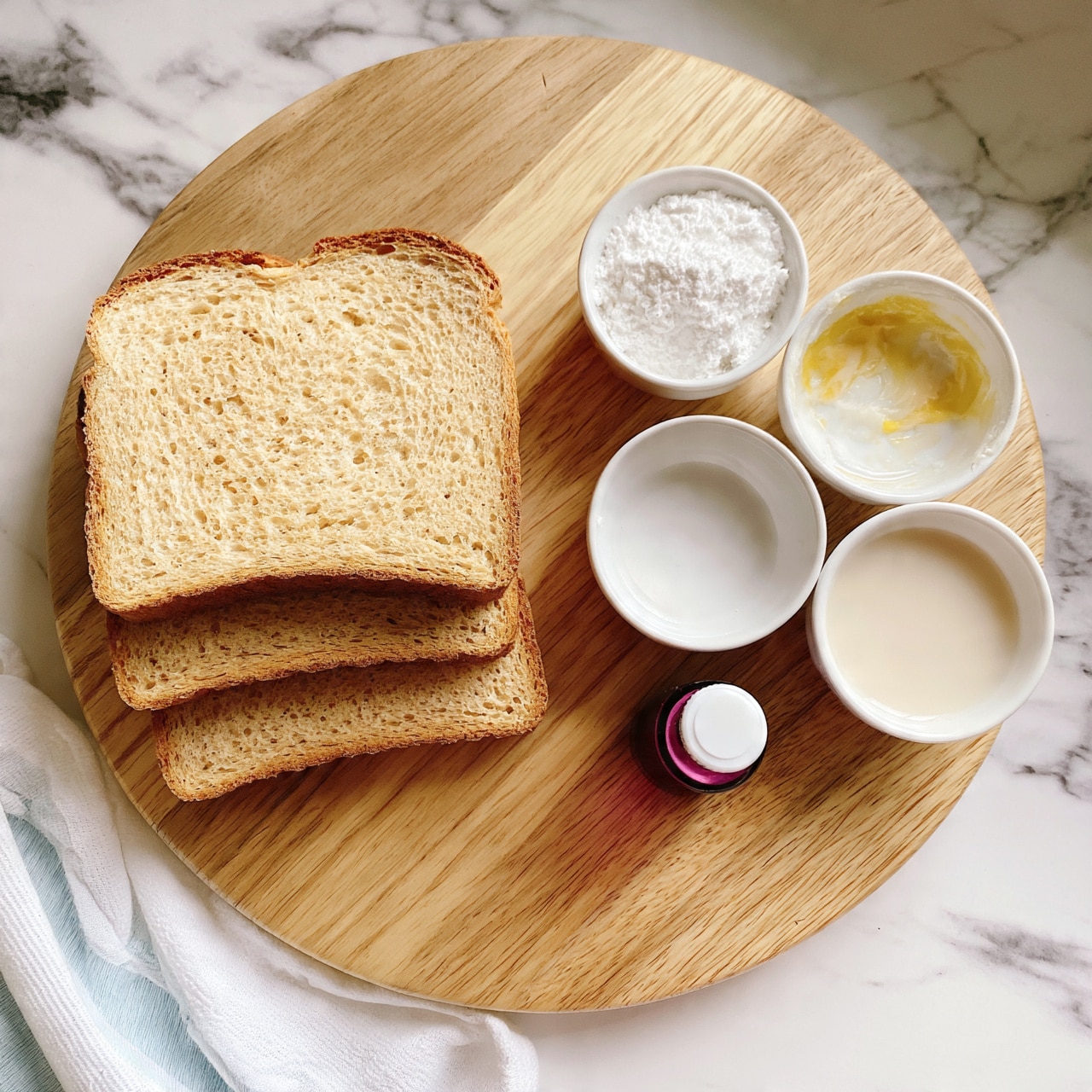 square image A round wooden board holding three slices of light brown bread stacked slightly overlapping on the left side; to the right, three small white bowls arranged in a triangle shape, one bowl has white powder, another has a yellow and white mixture, and the third contains a off-white liquid. In front of the bowls, there is a small dark pink bottle with a white cap. The set is placed on a white marbled surface with a white and light blue striped cloth visible on the bottom left side. Photo taken with an iphone --ar 4:5 --v 7
