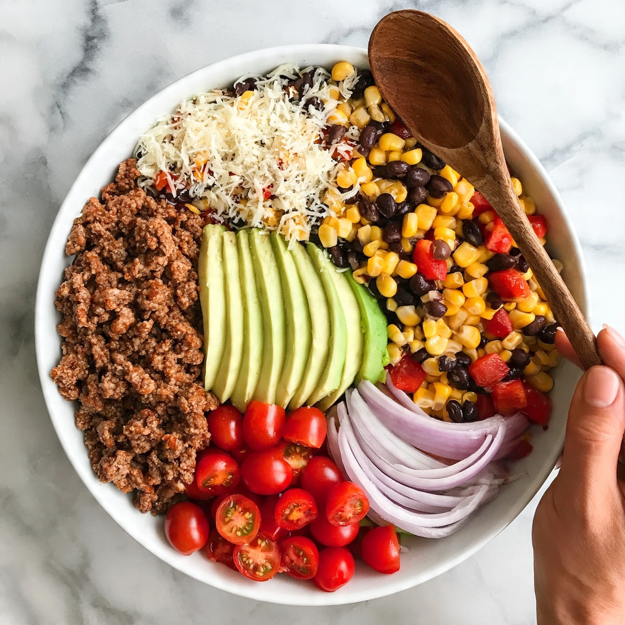 square image A white bowl filled with a colorful layered salad sits on a white marbled surface. The bowl contains several sections: in one part, there is a layer of cooked ground meat in brown color; next to it, sliced pale green avocado pieces are neatly placed upright. Another section shows halved small bright red cherry tomatoes. On the side, there are thin curved slices of light purple onions. At the top, there is a mix of yellow corn kernels, black beans, and diced red peppers, topped with a light layer of shredded white cheese. The bowl’s rim is visible, and a woman's hand holds a wooden spoon nearby. The photo taken with an iphone --ar 4:5 --v 7