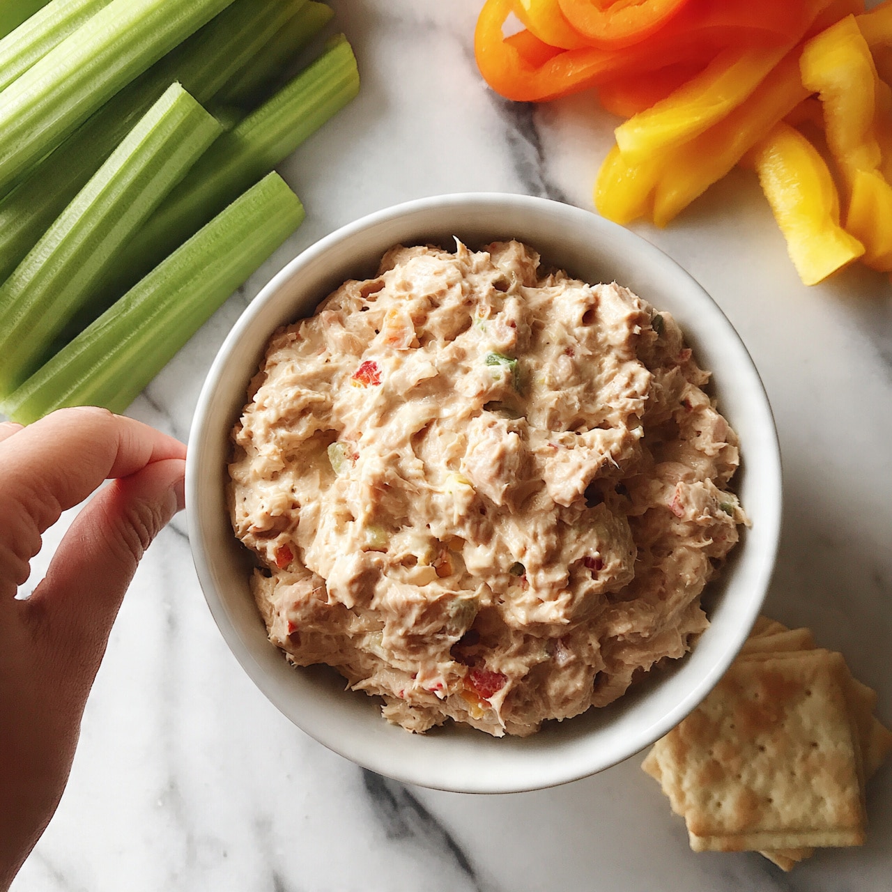 square image A white bowl filled with creamy, light brown tuna salad mixed with small red and green bits, placed at the center of a white marbled surface. Surrounding the bowl are bright green celery sticks on the left, vibrant orange and yellow bell pepper slices at the top right, and a small stack of square beige crackers at the bottom right. A woman's hand is reaching in from the left side toward the celery sticks. The scene is warmly lit and inviting. photo taken with an iphone --ar 4:5 --v 7