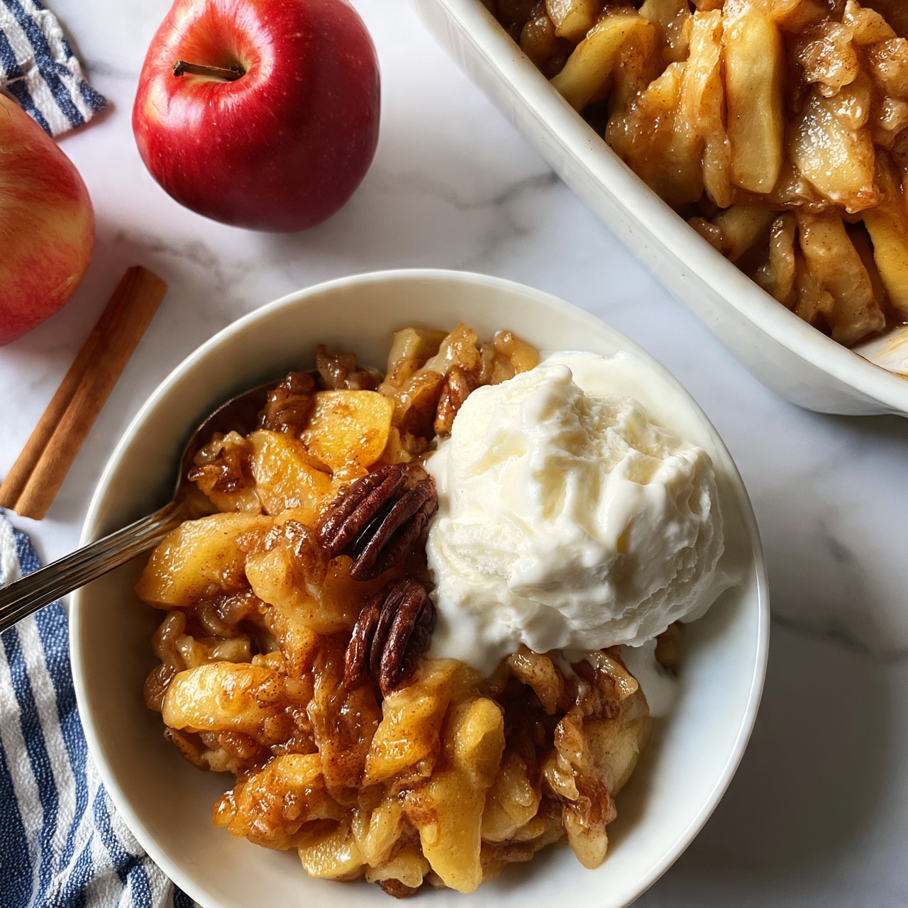 square image A white bowl filled with warm, cooked apple slices mixed with cinnamon and brown sugar, topped with a scoop of melting white vanilla ice cream that has a smooth texture, garnished with a few dark brown pecans. The apple mixture is golden brown with soft, glossy pieces and is placed at the bottom, while the ice cream scoop sits on the side, slightly melting into the apples. A spoon rests inside the bowl on the left side. Beside the bowl is a white baking dish with more of the apple mixture, showing layers of soft, caramelized apple slices. Two whole red apples and a cinnamon stick are placed on a white marbled surface next to the bowl, which also has a blue and white striped cloth nearby. photo taken with an iphone --ar 4:5 --v 7