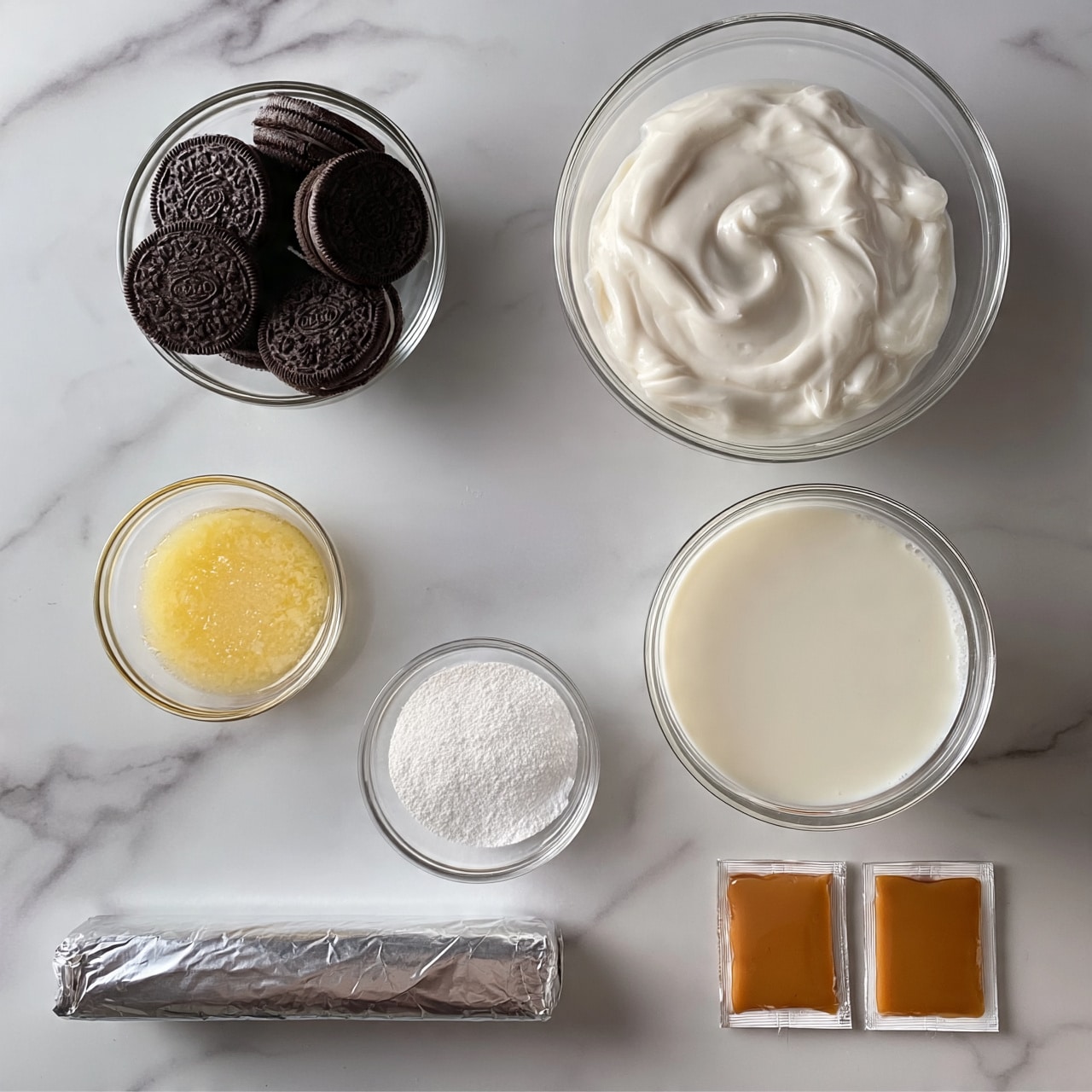 square image A top view of a white marbled surface with several clear glass bowls and packets arranged neatly: a small clear bowl filled with whole chocolate sandwich cookies on the top left, a large clear bowl filled with white cream on the top right, a medium clear bowl filled with milk below the cream, a small clear bowl with melted butter to the left of the milk, a small clear bowl with white powdered sugar beneath the butter, a stick of butter wrapped in silver foil on the bottom left, and two small caramel-colored packets side by side on the bottom right. photo taken with an iphone --ar 4:5 --v 7