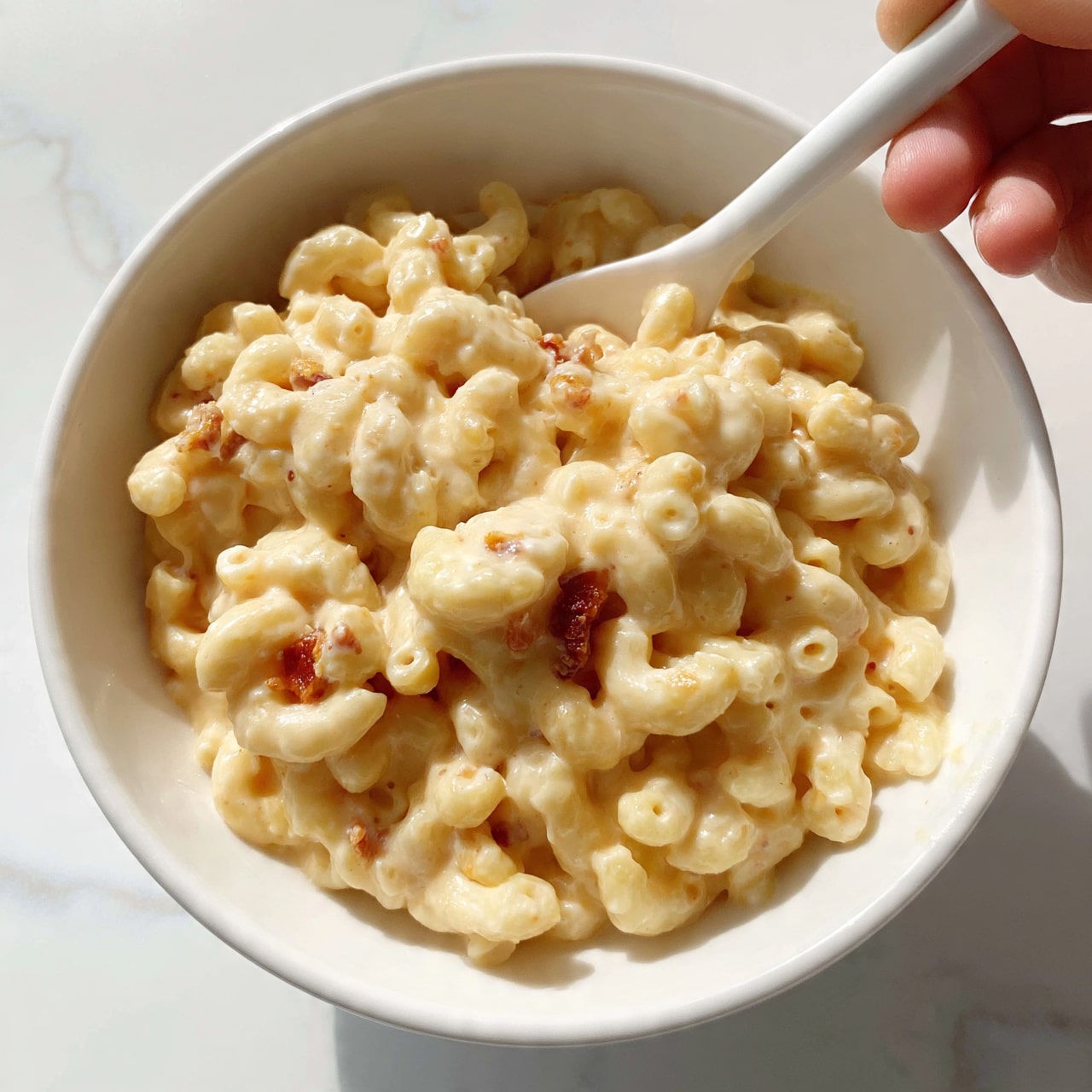 square image The image shows a close-up of creamy macaroni and cheese in a white bowl. The macaroni is light yellow in color with small bits of reddish ham mixed in. The cheese sauce looks thick and smooth, coating every piece of pasta evenly. A white spoon is placed inside the bowl with a woman's hand holding it from the top right side. The bowl sits on a white marbled surface, and the shot is bright and clear, focusing on the texture of the macaroni and cheese. photo taken with an iphone --ar 4:5 --v 7