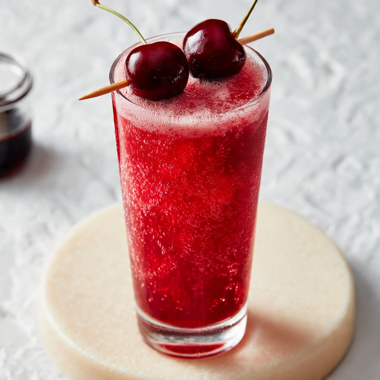 square image A tall, clear glass is filled with a bright red, bubbly drink that has a frothy layer on top. Two shiny dark red cherries with green stems are placed on a small wooden stick, resting across the rim of the glass. The glass is placed on a light cream round surface with a soft texture. In the background, there is a small glass jar with a dark liquid inside. The whole scene is set on a white marbled textured background. photo taken with an iphone --ar 4:5 --v 7