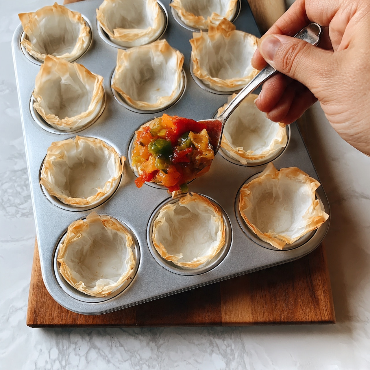 square image A close-up image of a silver muffin tray holding six white filo pastry cups, each with thin, light brown crispy edges. One cup in the tray is being filled with a mix of colorful vegetables: red, orange, and green pieces that appear soft and cooked, placed gently with a silver spoon. The tray sits on a wooden board, which is positioned on a white marbled texture surface. The scene shows a woman's hand holding the spoon while adding the vegetables inside the pastry cup photo taken with an iphone --ar 4:5 --v 7