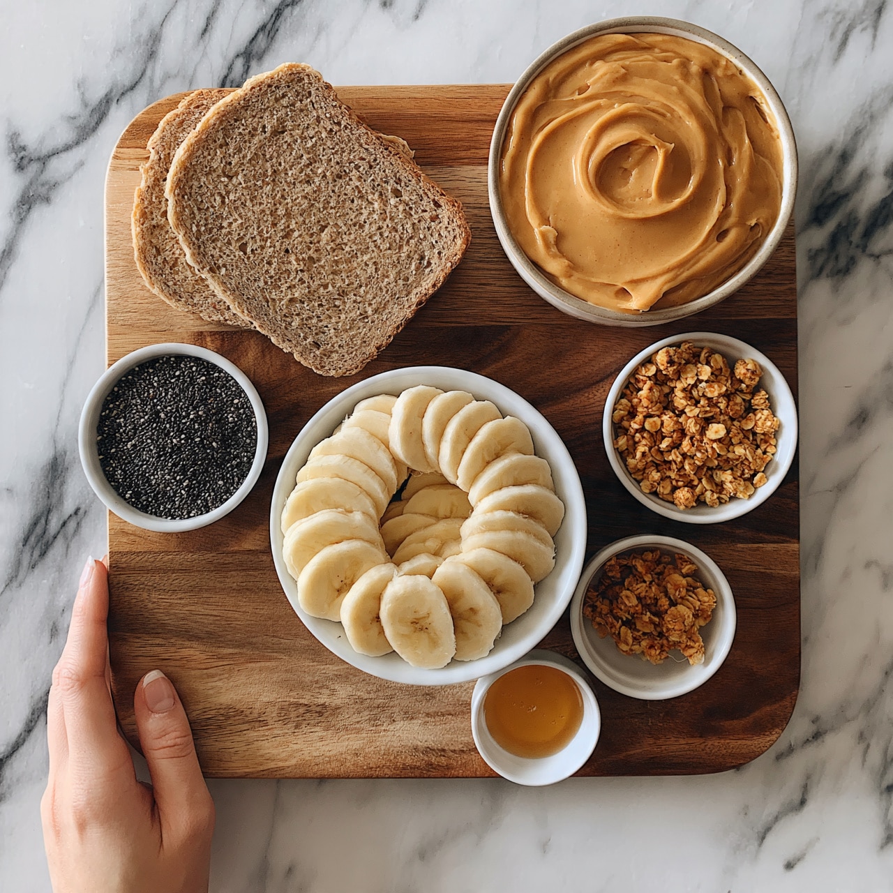 square image The image shows a wooden board set on a white marbled surface with two slices of brown bread placed slightly overlapping near the top left. To the top right, there is a round bowl filled with creamy peanut butter, swirled on top. Below the bread, a white bowl contains sliced bananas arranged neatly in a circle. Around the bowl, three small white bowls are arranged: one to the left filled with black chia seeds, one to the right filled with golden crunchy granola, and one below to the right with a small amount of honey. A woman's hand is gently holding the edge of the board. Photo taken with an iphone --ar 4:5 --v 7