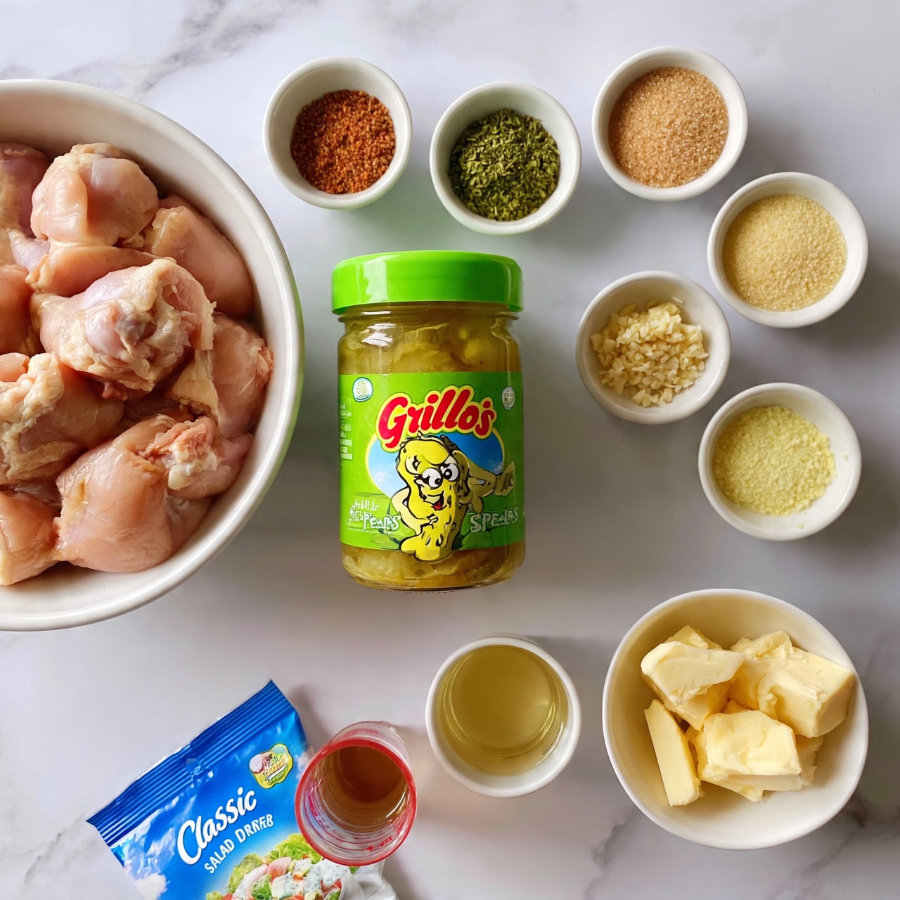 square image The image shows ingredients arranged neatly on a white marbled surface: a white bowl filled with raw pink chicken pieces sits on the left; next to it is a green jar of Grillo’s pickle spears with a bright green lid and a cartoon pickle on the label. Around the jar are small white bowls holding various spices and seasonings, including a reddish powder, green dried herbs, light brown sugar, finely chopped garlic, and some pale yellow powders. There is a small cup of clear golden liquid and a white bowl with chunks of pale yellow butter. Near the bottom left, a blue packet of classic ranch salad dressing mix and a small transparent red measuring cup with a brown liquid inside complete the layout. Photo taken with an iphone --ar 4:5 --v 7