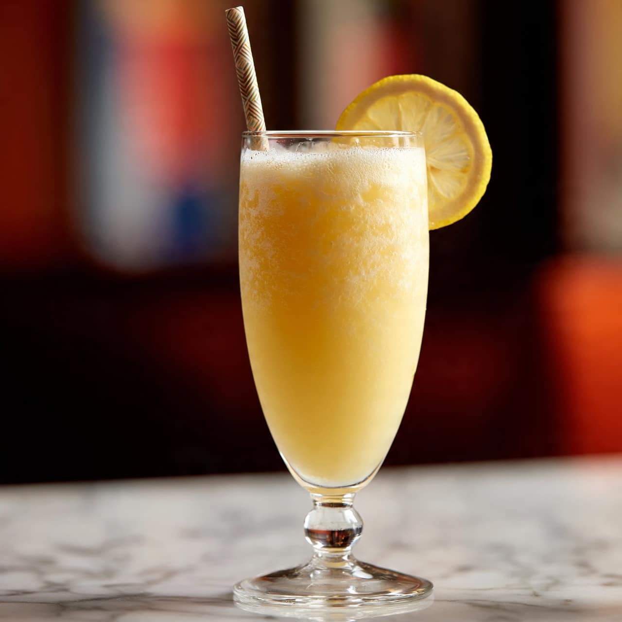 square image A tall clear glass with a short stem filled with a light yellow drink that looks smooth and slightly foamy at the top, with a patterned straw standing upright in the glass. A thin, fresh lemon slice is placed on the edge of the glass near the straw. The background is blurry with warm colors, and the glass is set on a dark wooden surface, replaced in the description with a white marbled texture. Photo taken with an iphone --ar 4:5 --v 7