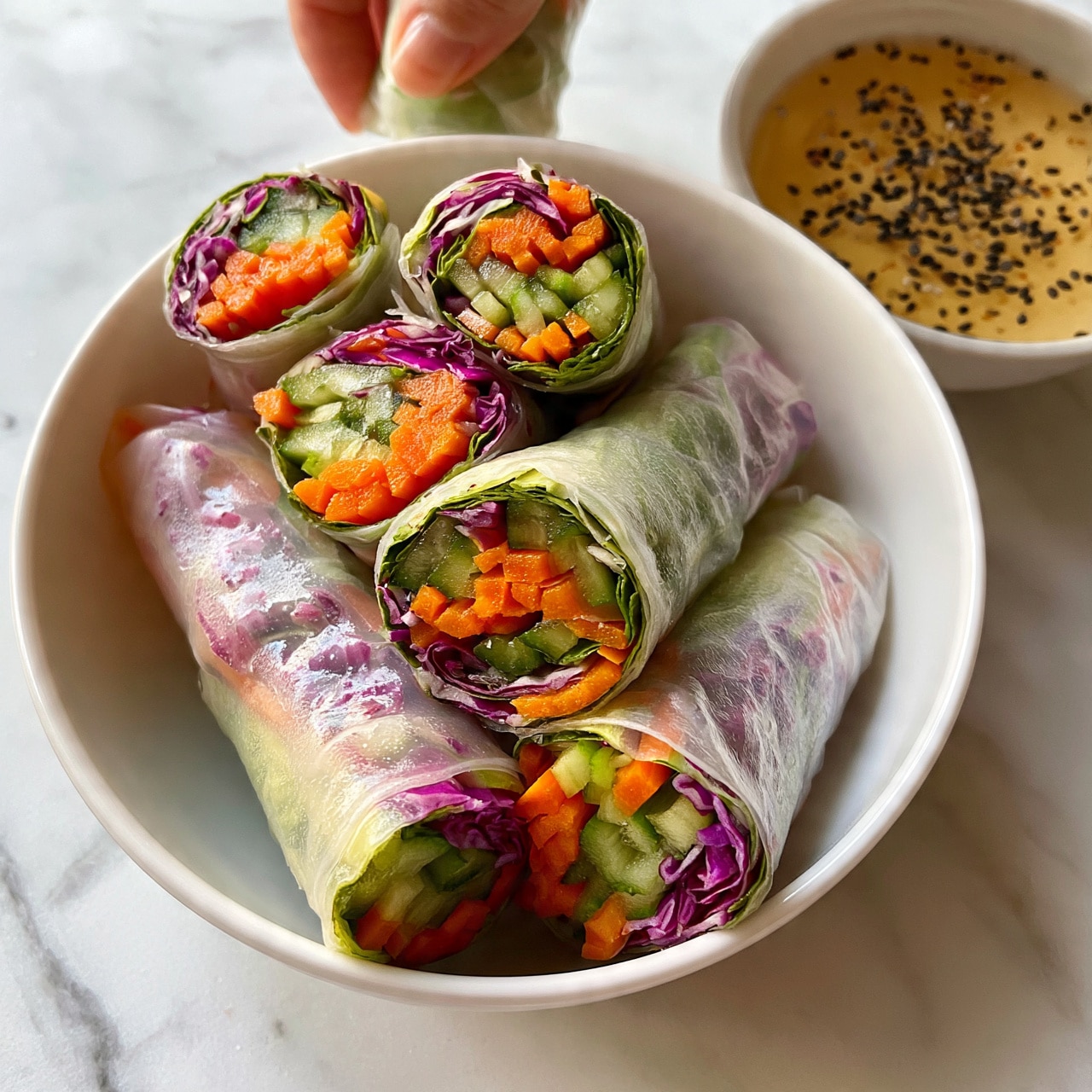 square image The image shows six fresh spring rolls arranged neatly in a white bowl, each roll layered with bright orange carrot strips, green cucumber slices, and purple cabbage, wrapped in translucent rice paper. The rolls have a slightly shiny texture, showing the colorful vegetables inside. On the right side of the bowl, there is a small white bowl filled with light yellow dipping sauce sprinkled with black sesame seeds. The background features a clean white marbled surface. A woman's hand is partially visible holding one spring roll above the bowl. photo taken with an iphone --ar 4:5 --v 7