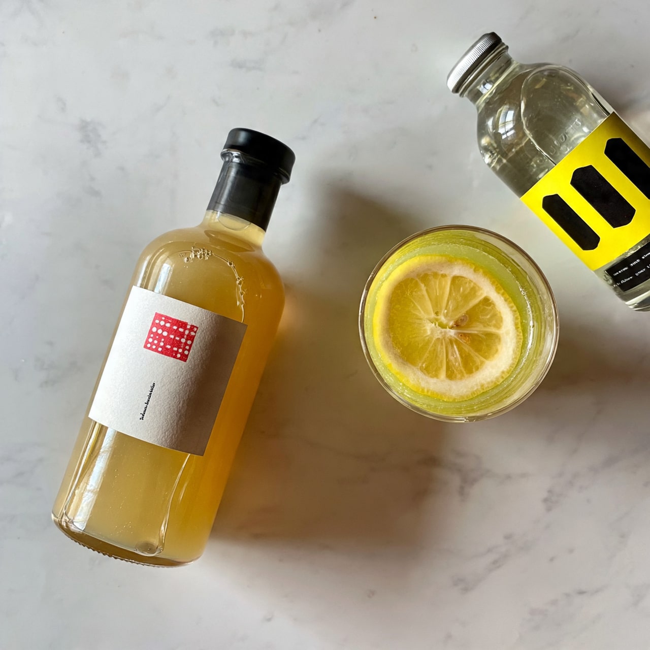 square image A light brown bottle with a white label and red text is placed on a white marbled surface, slightly tilted to the left. Next to it, on the right side, is a small clear glass with a yellow drink inside and a slice of lemon floating on top. Further to the right, there is a transparent bottle with a yellow and black label. All items are spaced out evenly on the white marbled surface. photo taken with an iphone --ar 4:5 --v 7