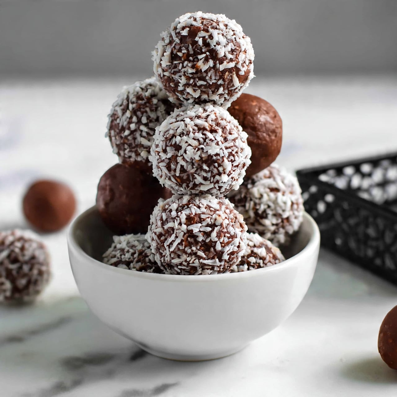 square image A small white bowl filled with round chocolate balls, some fully covered in shredded white coconut and others plain chocolate, stacked in a pyramid shape. The bowl sits on a white marbled surface with a few chocolate balls scattered around it. To the side, a black decorative rectangular dish is partially visible. The texture of the chocolate balls looks smooth and slightly rough where the coconut flakes stick. The photo taken with an iphone --ar 4:5 --v 7