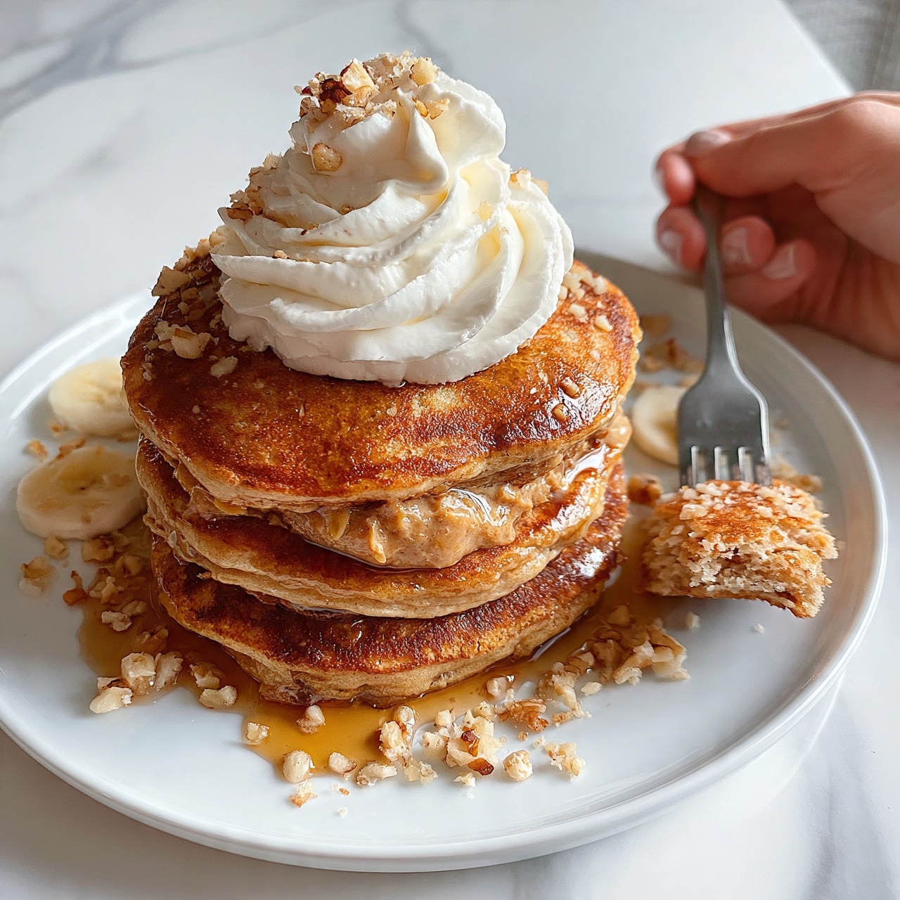 square image The image shows a white plate on a white marbled surface, holding a stack of three thick pancakes. The pancakes are golden brown with a slightly rough texture. Between the layers, there is a spread of light brown nut butter. On top of the stack, there is a generous swirl of white whipped cream. Around the plate, especially near the pancakes, are small banana slices and sprinkled chopped walnuts. A woman's hand holds a fork resting on the right side of the plate with a piece of pancake on it. The syrup glistens on the pancakes and pools gently at the base. photo taken with an iphone --ar 4:5 --v 7