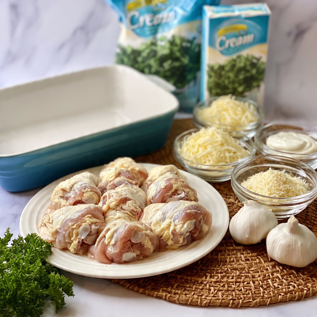 square image The image shows raw chicken pieces placed on a white round plate in front of a white baking dish with a blue outside. To the right of the chicken, there are several small clear glass bowls filled with shredded cheese, a white creamy ingredient, and grated cheese, arranged neatly on a woven brown placemat. Behind the chicken and bowls, a bag of chopped spinach and a box of cream cheese are standing upright. Two garlic bulbs sit next to the bowls, along with a sprig of fresh green parsley in the lower left corner. The background is a white marbled texture. Photo taken with an iphone --ar 4:5 --v 7