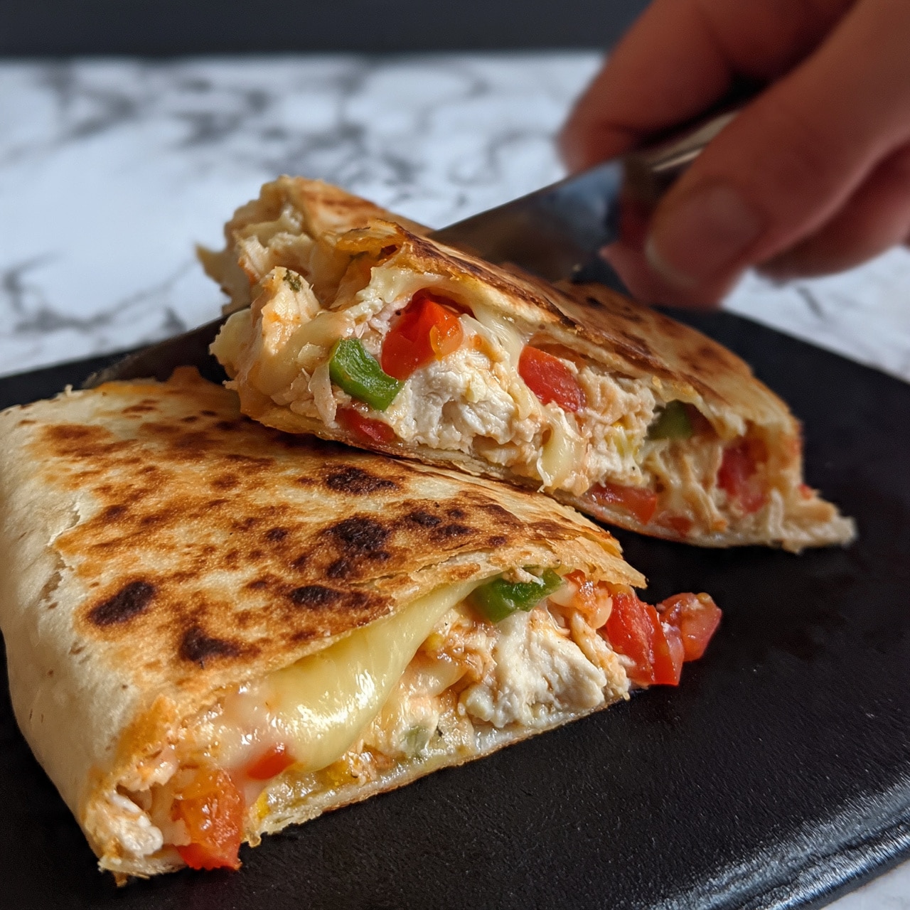 square image The image shows two triangular slices of quesadilla on a black surface with a white marbled background. The quesadilla has a golden-brown, slightly crispy outer layer with visible brown spots. Inside, there is a colorful mix of melted cheese that is soft and light yellow, diced red tomatoes, green bell peppers, and chunks of creamy white chicken. A woman’s hand is holding a knife and slicing through one of the quesadilla pieces, with careful, precise motion. Photo taken with an iphone --ar 4:5 --v 7