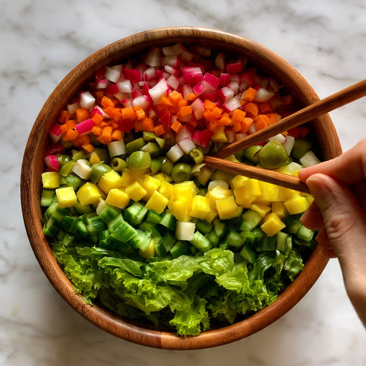 square image A wooden bowl filled with a fresh salad showing about four layers: the bottom layer is bright green leafy lettuce, followed by chopped green cucumbers and yellow cubes of cheese, next are small green olives and white cubes, and on top are small pieces of red radish and orange chopped carrots scattered evenly. Two wooden chopsticks rest inside the bowl, with a woman's hand slightly touching them. The bowl sits on a white marbled surface. Photo taken with an iphone --ar 4:5 --v 7