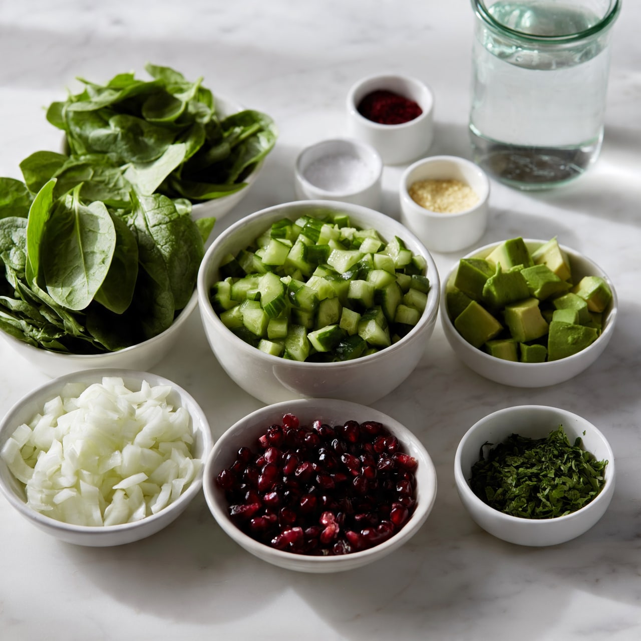 square image The image shows multiple white bowls and small white containers placed on a white marbled surface. From left to right, there is a bowl filled with fresh green spinach leaves, another bowl with smaller spinach leaves behind it, a medium-sized bowl full of chopped green cucumbers, and a small bowl with cubed green avocado pieces. In front, there is a bowl of chopped white onions, a bowl with dark red pomegranate seeds, and a small bowl with chopped green herbs. There are also three small white containers holding a dark red powder, some salt, and a light yellow powder, and a glass jar filled with clear water. Everything is cleanly arranged and well-lit, photo taken with an iphone --ar 4:5 --v 7