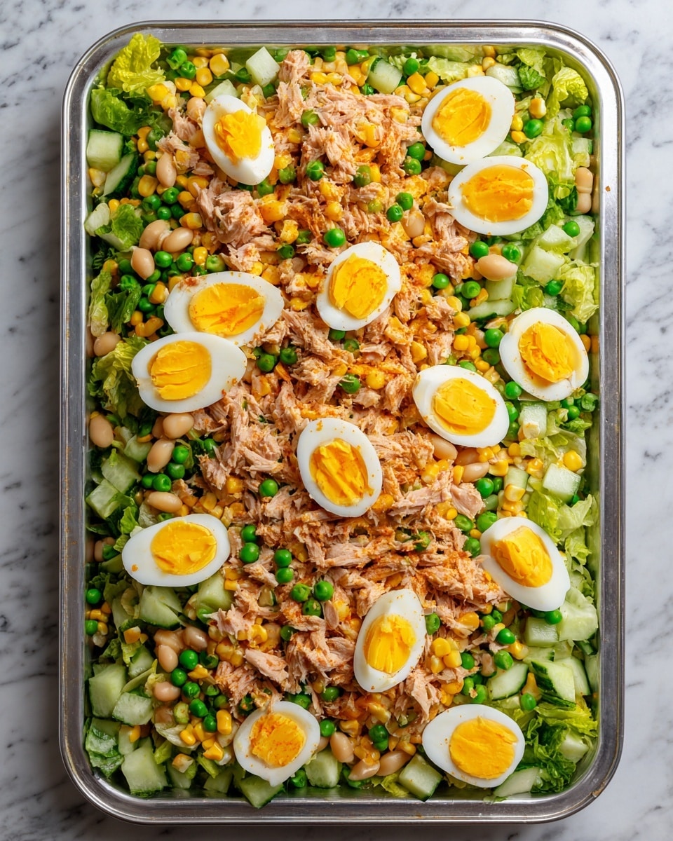A large rectangular aluminum tray filled with a colorful layered salad resting on a white marbled surface. The bottom layer consists of chopped green lettuce and sliced cucumber pieces, creating a fresh green base. Above this, there is a layer of white beans mixed with orange corn kernels scattered evenly. Small bright green peas are mixed in, adding more color and texture. On top, crumbled light pink tuna is spread evenly across the layers. The dish is topped with halved boiled eggs showing bright yellow yolks and smooth white edges, placed evenly around the surface. photo taken with an iphone --ar 4:5 --v 7