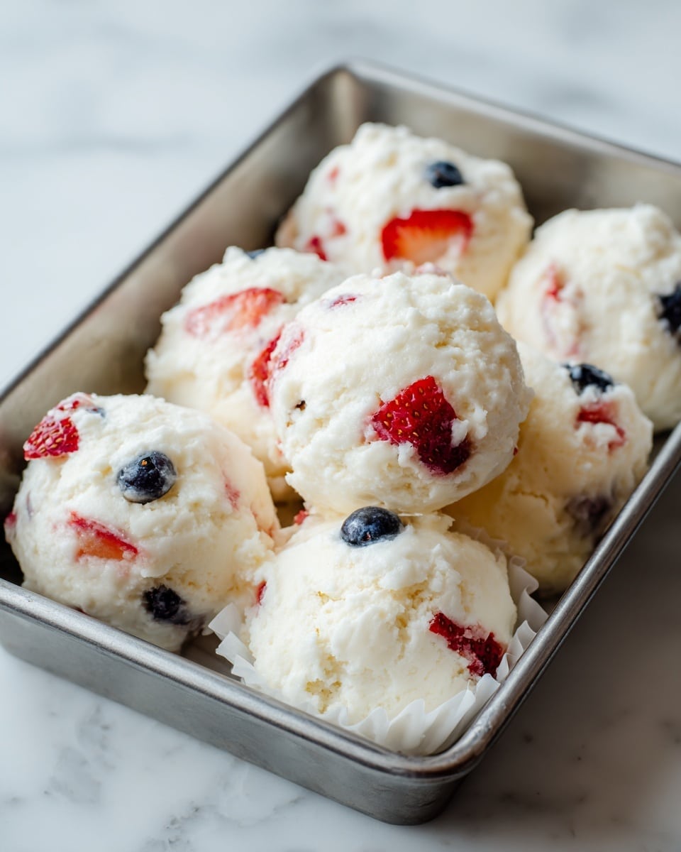 The image shows a metal tray filled with white frozen clusters that look soft and fluffy. Each cluster contains visible red pieces of strawberry and small blue blueberries mixed gently into the white creamy layer, giving it a colorful and fresh look. The texture looks smooth and slightly uneven, with the fruit parts sitting slightly inside the white layer, creating a natural blend. The clusters have a rough round shape with some showing a scalloped edge from a white paper liner. The tray is placed on a white marbled surface, enhancing the clean and fresh appearance of the dish. Photo taken with an iphone --ar 4:5 --v 7