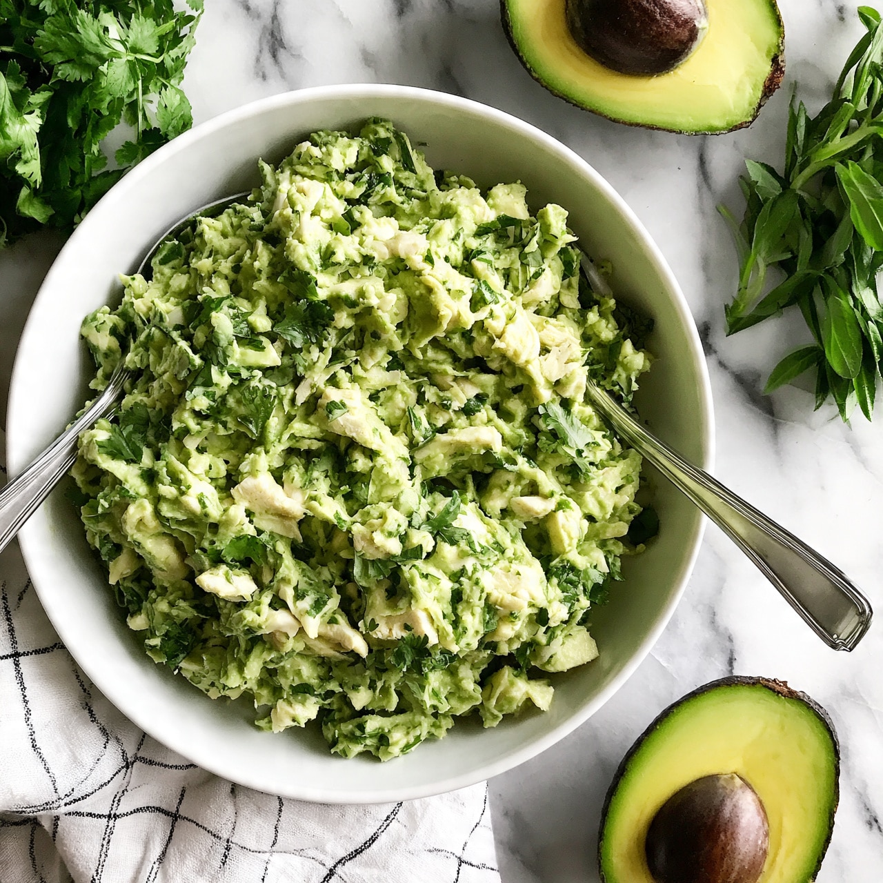 square image A white bowl filled with a chunky green mixture that looks like mashed avocado mixed with small pieces of white chicken and green herbs. The mix is unevenly textured with some larger chunks and bits of cilantro spread throughout. Two silver spoons are placed on opposite sides inside the bowl. The bowl sits on a white marbled surface with a halved avocado showing its dark brown seed and bright green flesh nearby. To the side, fresh green leafy herbs are visible, and a white cloth with a black grid pattern is laid under part of the bowl. Photo taken with an iphone --ar 4:5 --v 7 square image