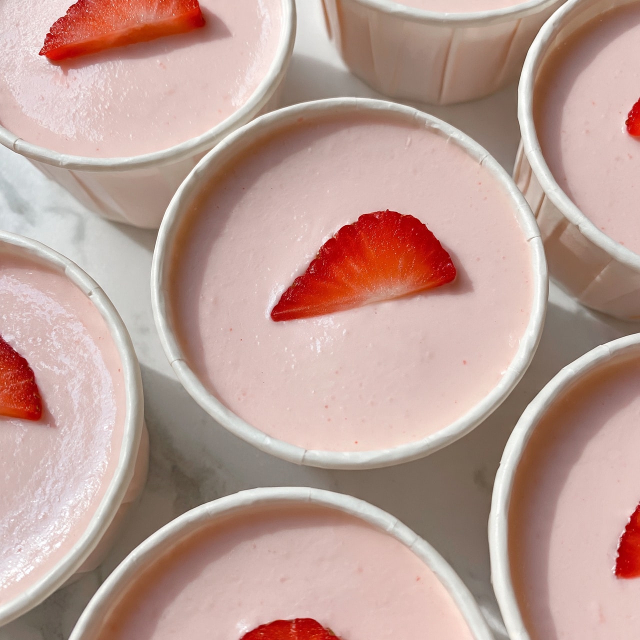 square image The image shows several small white paper cups filled with light pink creamy dessert that has a smooth, soft texture. Each dessert is topped with a thin, bright red slice of strawberry placed in the center. The desserts are arranged closely together on a white marbled surface, creating a neat pattern. The light pink cream and the red strawberry slices add a fresh, delicate look to the simple, sweet treats. photo taken with an iphone --ar 4:5 --v 7