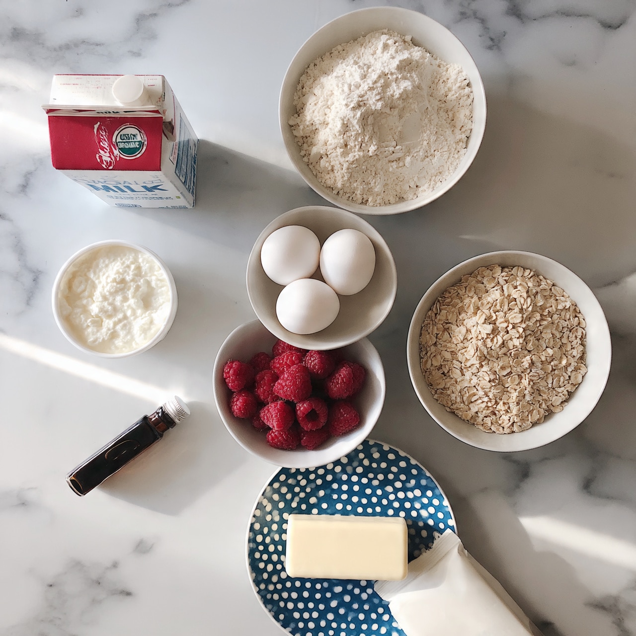 square image The image shows a white marbled surface with several bowls and packages arranged neatly. There are three white eggs in a white bowl near the center. A white bowl with fresh red raspberries sits below the eggs, and to the right, a white bowl is filled with light brown rolled oats. Above the eggs, a white bowl holds a heap of white flour. To the left are a carton of organic milk with red and white colors, a white container of cottage cheese, and a small dark bottle with vanilla extract lying horizontally. In the lower center, there's a blue and white polka-dotted plate holding a rectangular stick of butter. At the bottom right, a white package of white chocolate is placed partially on the plate. photo taken with an iphone --ar 4:5 --v 7