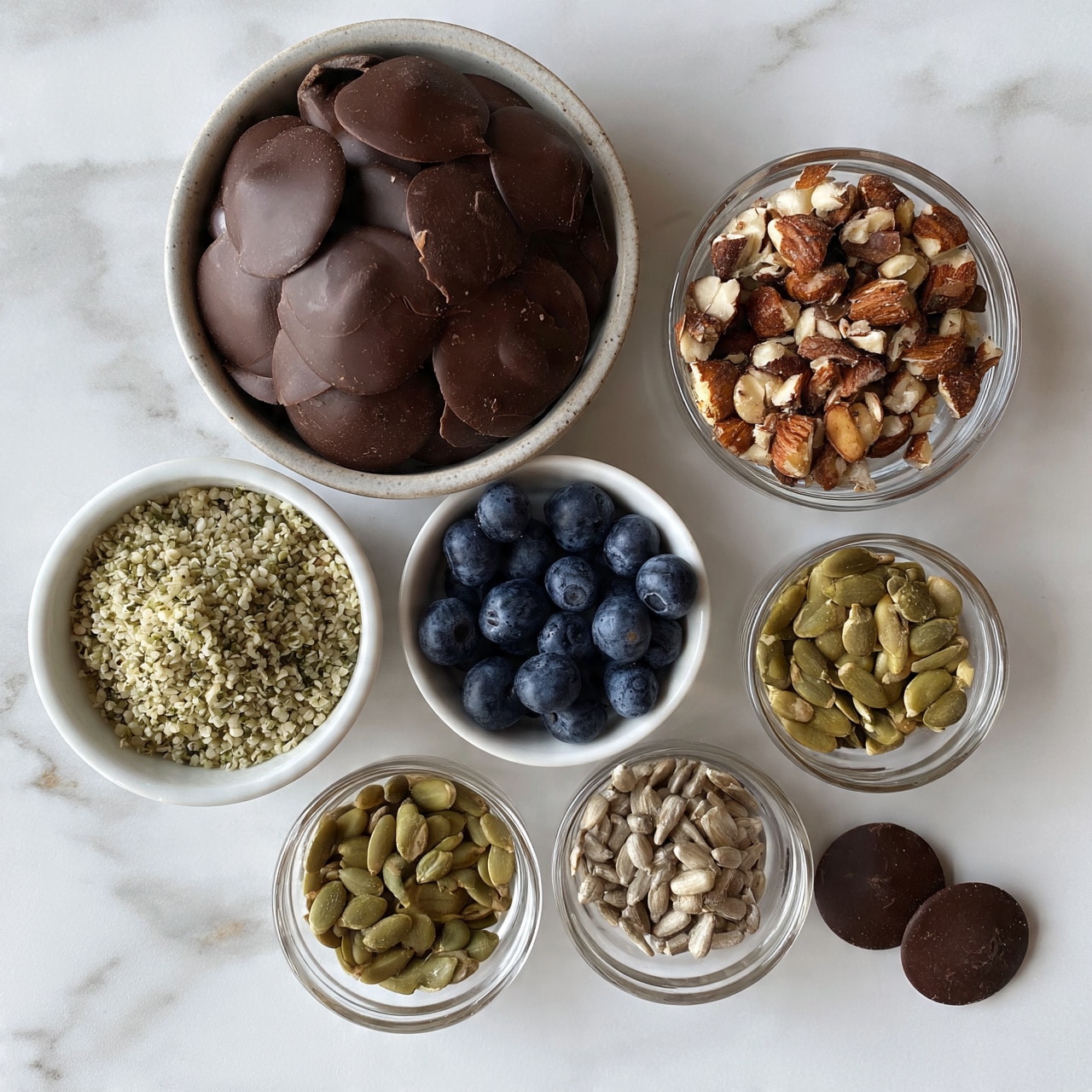 square image Six small bowls with different ingredients are placed on a white marbled surface. The largest bowl contains smooth, dark chocolate discs. Another bowl holds round, plump blueberries that are deep blue in color. There is a small white bowl filled with light greenish hemp seeds. Next to it is a clear bowl filled with mixed nuts, mostly almonds, with a rough and chopped texture. Two smaller clear bowls contain pumpkin seeds and sunflower seeds, both with a dry and slightly shiny texture. A few dark chocolate discs are scattered on the surface near the bowls. The photo was taken with an iphone --ar 4:5 --v 7