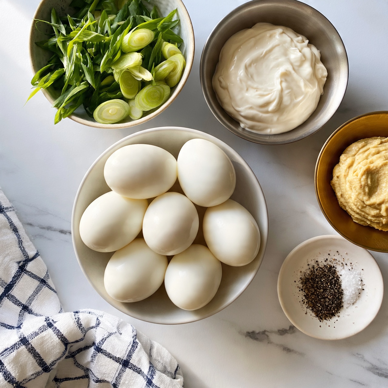 square image Five peeled hard boiled eggs sit tightly together in a white bowl on a white marbled surface, next to a bowl of sliced green onions with bright green leaves and pale green rings, a metal bowl with smooth white mayonnaise, a brass bowl holding creamy mustard, and a small white plate with coarse black pepper and salt. A white and navy checkered cloth is softly folded in the bottom left corner. The light is soft and natural, showing clear textures of all ingredients. Photo taken with an iphone --ar 4:5 --v 7