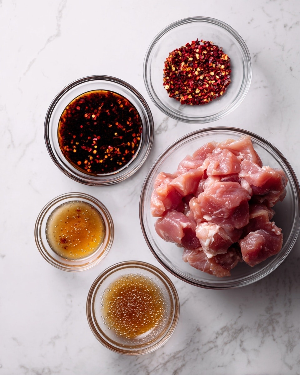 The image shows five clear glass bowls arranged on a white marbled surface. The largest bowl at the bottom right contains raw pink chicken pieces cut into chunks. Above this, a medium bowl holds a dark brown liquid with red pepper flakes floating on top. To the right side of that bowl is a smaller bowl with a dark, almost black liquid. Below the chili sauce bowl, another small bowl contains a light golden liquid with small bubbles on its surface. At the top center is the smallest bowl holding red pepper flakes. All bowls are transparent and the setup is clean and minimal. Photo taken with an iphone --ar 4:5 --v 7