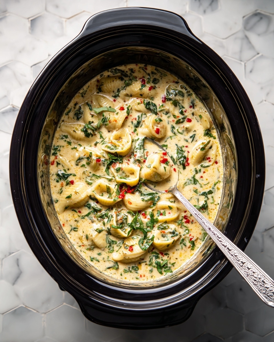 The image shows a black slow cooker filled with a creamy soup that has several layers of small pasta pieces shaped like stuffed shells, mixed with green leafy herbs and small red bits, possibly peppers or tomatoes. The soup broth has a light yellow color with a slightly oily texture, and the herbs and red bits are evenly spread throughout the soup. A silver spoon with a patterned handle rests inside the cooker, partially submerged. The slow cooker sits on a white marbled surface with a soft hexagonal pattern in the blurred background, creating a clean and bright setting photo taken with an iphone --ar 4:5 --v 7