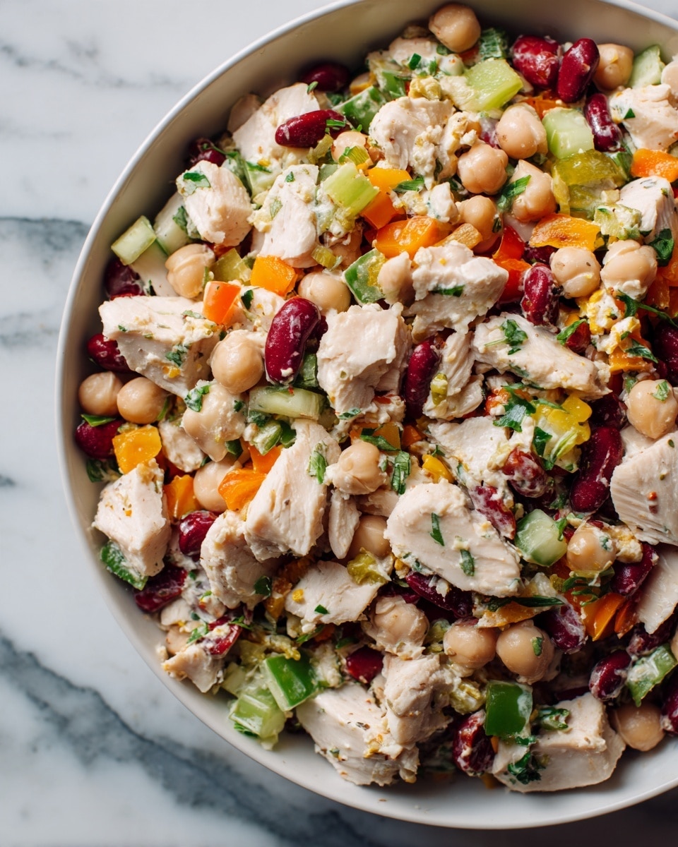 The image shows a close-up of a colorful mixed bean and chicken salad in a white bowl, sitting on a white marbled surface. The salad has several layers and textures: large chunks of white cooked chicken scattered across the top, mixed with light beige chickpeas, bright red kidney beans, and white cannellini beans. There are pieces of orange bell pepper and green celery slices mixed evenly throughout, with small finely chopped green herbs sprinkled on top, adding fresh color. The salad looks fresh and mixed well, with all ingredients visible in similar amounts. Photo taken with an iphone --ar 4:5 --v 7