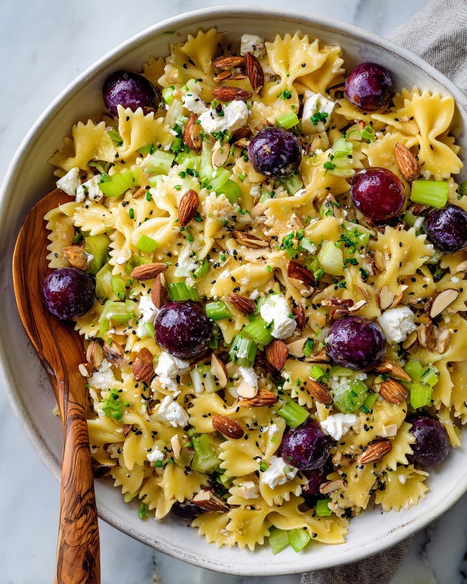 A close-up view of a large, shallow white bowl filled with a colorful pasta salad layered with small farfalle pasta pieces that are yellow and soft textured as the base layer, mixed with halved purple grapes scattered evenly throughout. On top of this are bright green chopped celery pieces adding crunch and freshness, along with small white cubes of cheese dispersed around. Thinly sliced almonds and whole almonds are spread on the surface for extra texture, and small black poppy seeds are sprinkled all over. The salad is garnished with finely chopped green onions, creating a fresh green contrast. A wooden spoon is resting on the edge of the bowl, all set on a white marbled surface. Photo taken with an iphone --ar 4:5 --v 7
