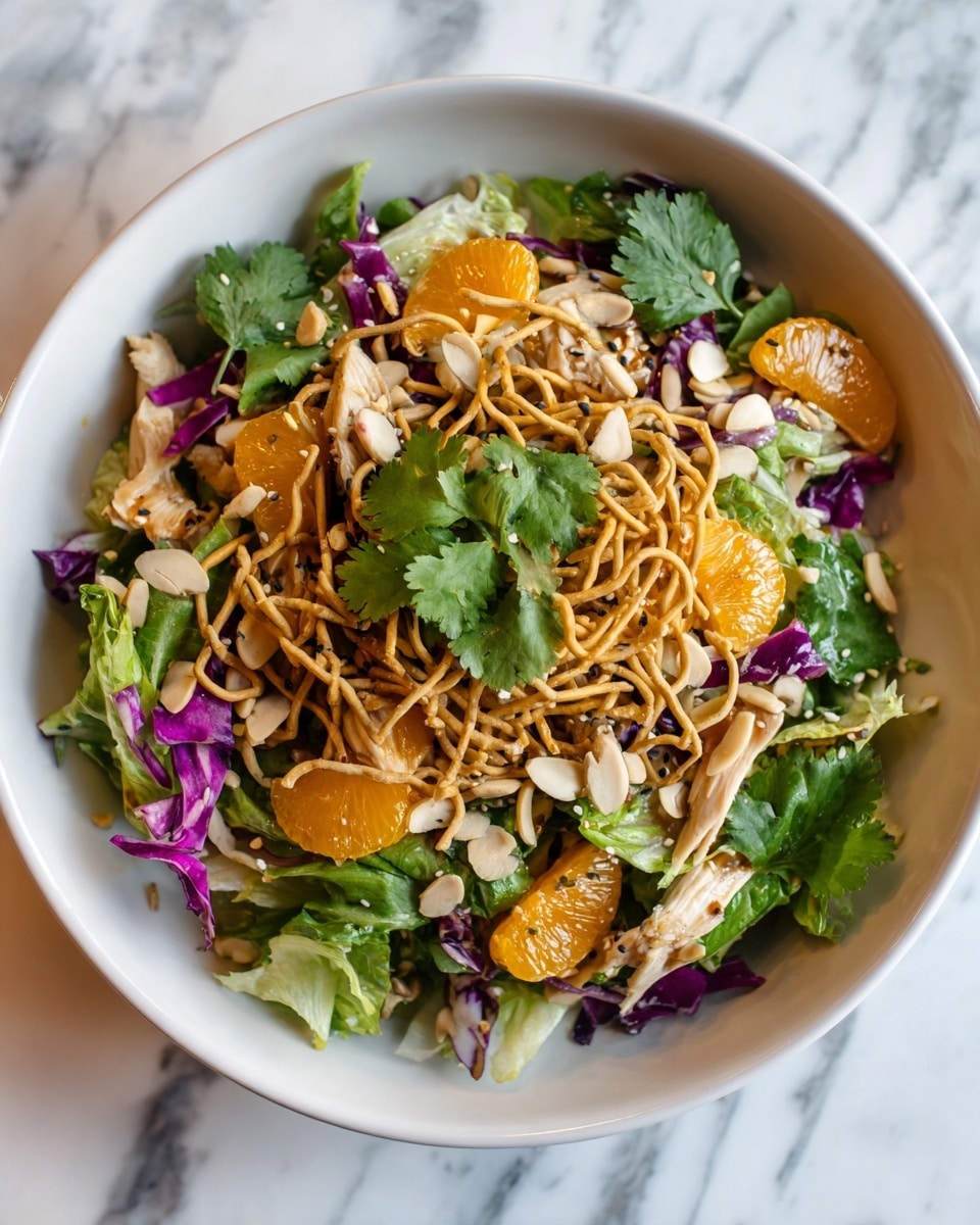 A close-up view of a salad served in a white bowl, resting on a white marbled surface. The salad has a base layer of mixed green lettuce and purple cabbage, making a vibrant green and purple bed. On top, tender pieces of light beige cooked chicken are scattered, blending with bright orange mandarin segments. The upper layer has crunchy, golden-brown fried noodle strips placed unevenly over the salad. Fresh green cilantro leaves and light-colored almond slices are sprinkled throughout, with some sesame seeds adding tiny white accents. The layers show a mix of fresh, crunchy, and soft textures in a colorful arrangement. Photo taken with an iphone --ar 4:5 --v 7