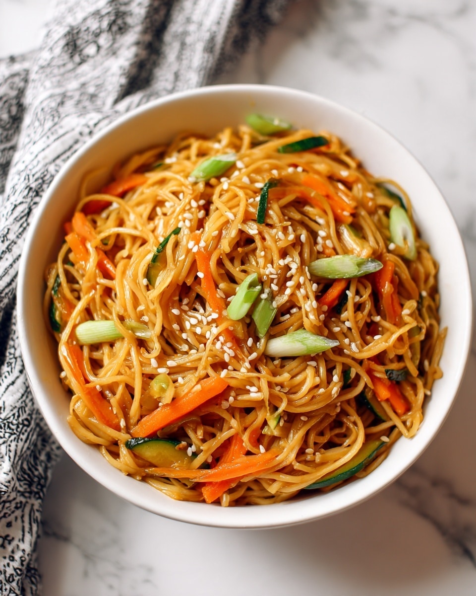 The image shows a white bowl filled with a mix of thin, golden-brown noodles and long, thin strips of orange carrots and green zucchini. The noodles and vegetables are coated lightly in a glossy sauce, making them look shiny and smooth. Small white sesame seeds are scattered evenly on top, along with slices of green onions that add a fresh look. The bowl is set on a white marbled surface, and a corner of a black and white cloth is visible in the background. photo taken with an iphone --ar 4:5 --v 7