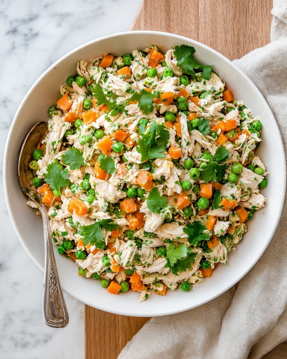 A large white bowl filled with a mixed salad containing light pink pieces of shredded chicken, small bright orange carrot cubes, and green peas spread evenly throughout. Fresh green cilantro leaves are scattered on top and mixed inside, giving a vibrant contrast in color. A silver spoon is resting inside the bowl on the left side, partially buried in the salad. The bowl sits on a light wooden surface with a beige cloth cloth to the right side, and the background has a clean white marbled texture. photo taken with an iphone --ar 4:5 --v 7