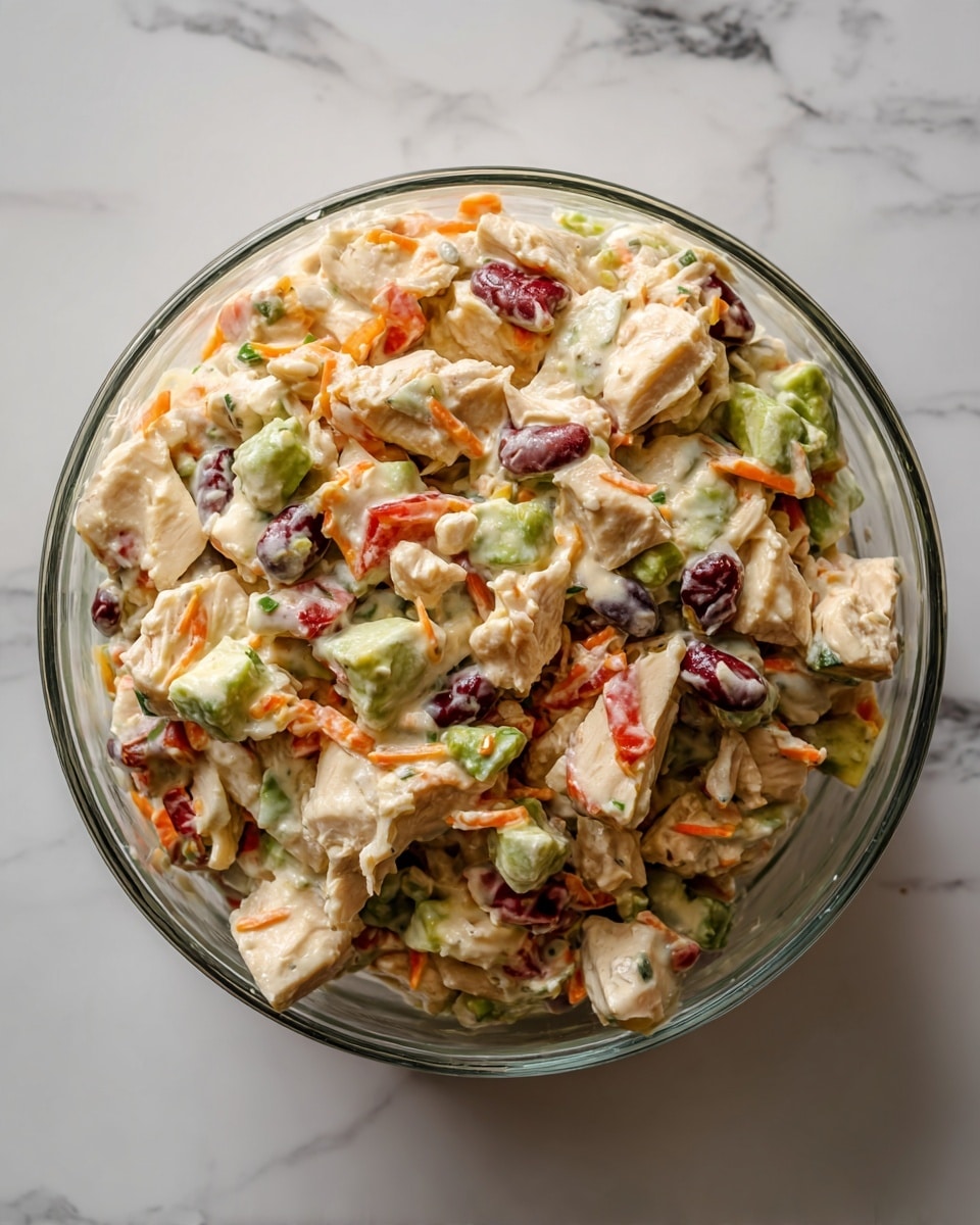 This image shows a clear glass bowl filled with a colorful chicken salad. The salad has several layers, starting with chunks of cooked chicken that are light brown with some white pieces. Mixed in are green avocado pieces, bright red tomato bits, and small black beans. There are also light orange shredded carrots throughout the dish. The salad looks creamy with some dressing lightly coating the ingredients. The bowl sits on a white marbled surface. photo taken with an iphone --ar 4:5 --v 7
