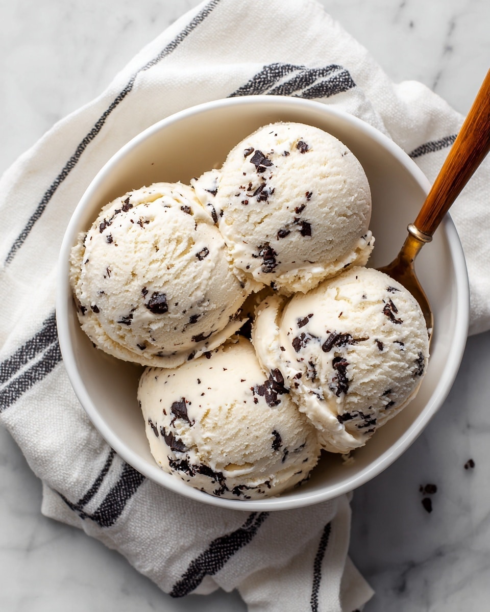 A white bowl filled with four scoops of light cream-colored ice cream with visible dark chocolate chips mixed throughout. The texture of the ice cream looks soft and creamy, and a spoon with a wooden handle is partially visible inside the bowl. The bowl is placed on a white marbled surface with a white cloth that has thin black stripes underneath it. photo taken with an iphone --ar 4:5 --v 7
