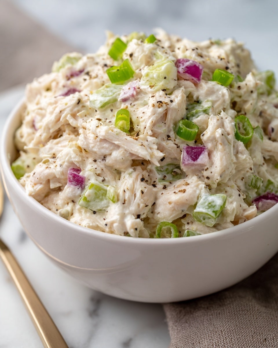 A white bowl filled with creamy chicken salad that has a thick texture with visible shredded white chicken pieces mixed with mayonnaise. Small chunks of pale green celery and tiny pieces of purple-red onions are scattered throughout, along with some green onion slices, adding small pops of color. The surface is dotted with specks of black pepper, giving a slight contrast, all placed on a white marbled surface photo taken with an iphone --ar 4:5 --v 7