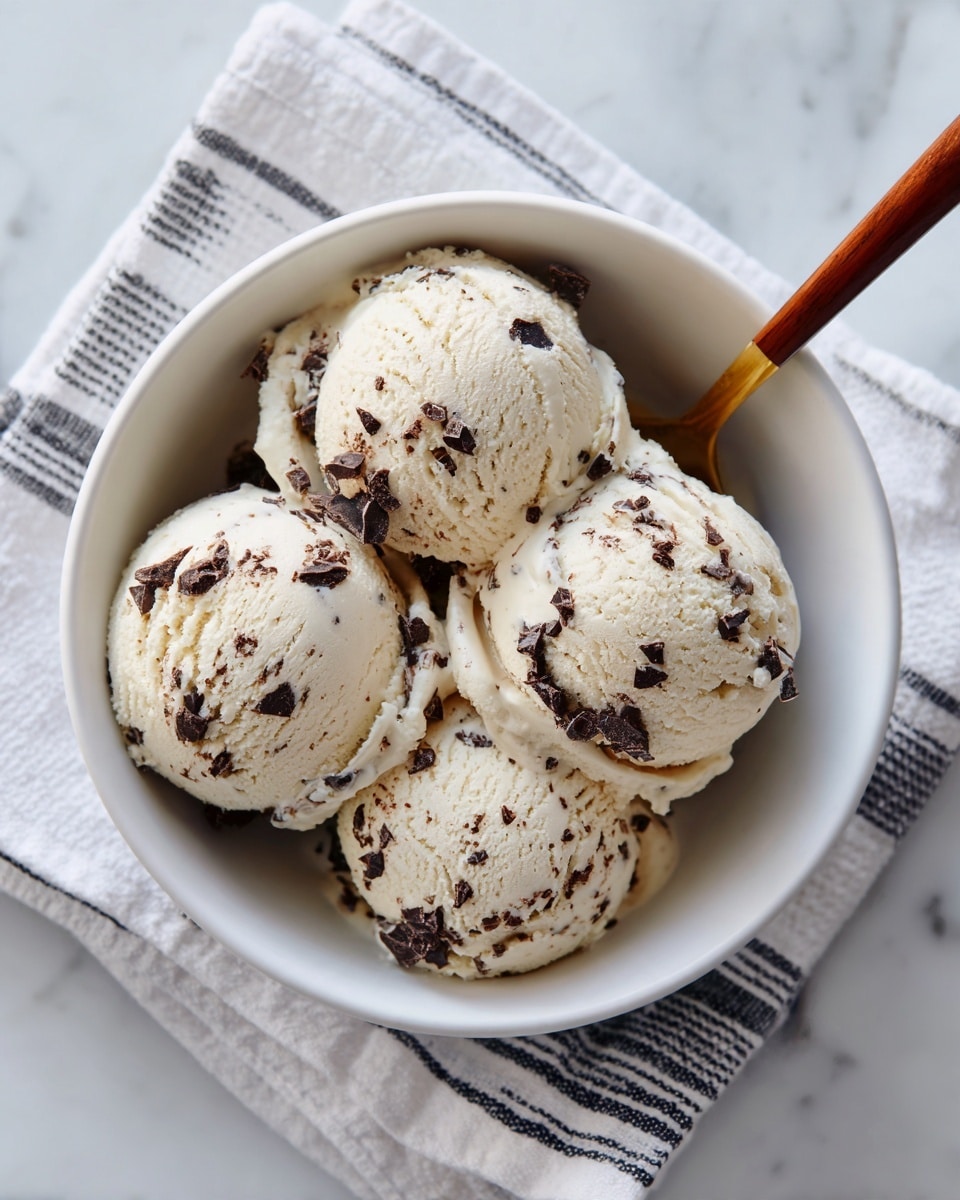 The image shows a white bowl filled with four scoops of creamy ice cream with chocolate chips mixed in. The ice cream is light beige in color with small dark brown chocolate chips scattered unevenly throughout each scoop. A spoon with a wooden handle is resting inside the bowl, its tip partly under the scoops. The bowl is placed on a white cloth with thin dark stripes, all set on a white marbled surface. The overall look is clean and simple, highlighting the smooth texture and soft swirls of the ice cream. photo taken with an iphone --ar 4:5 --v 7