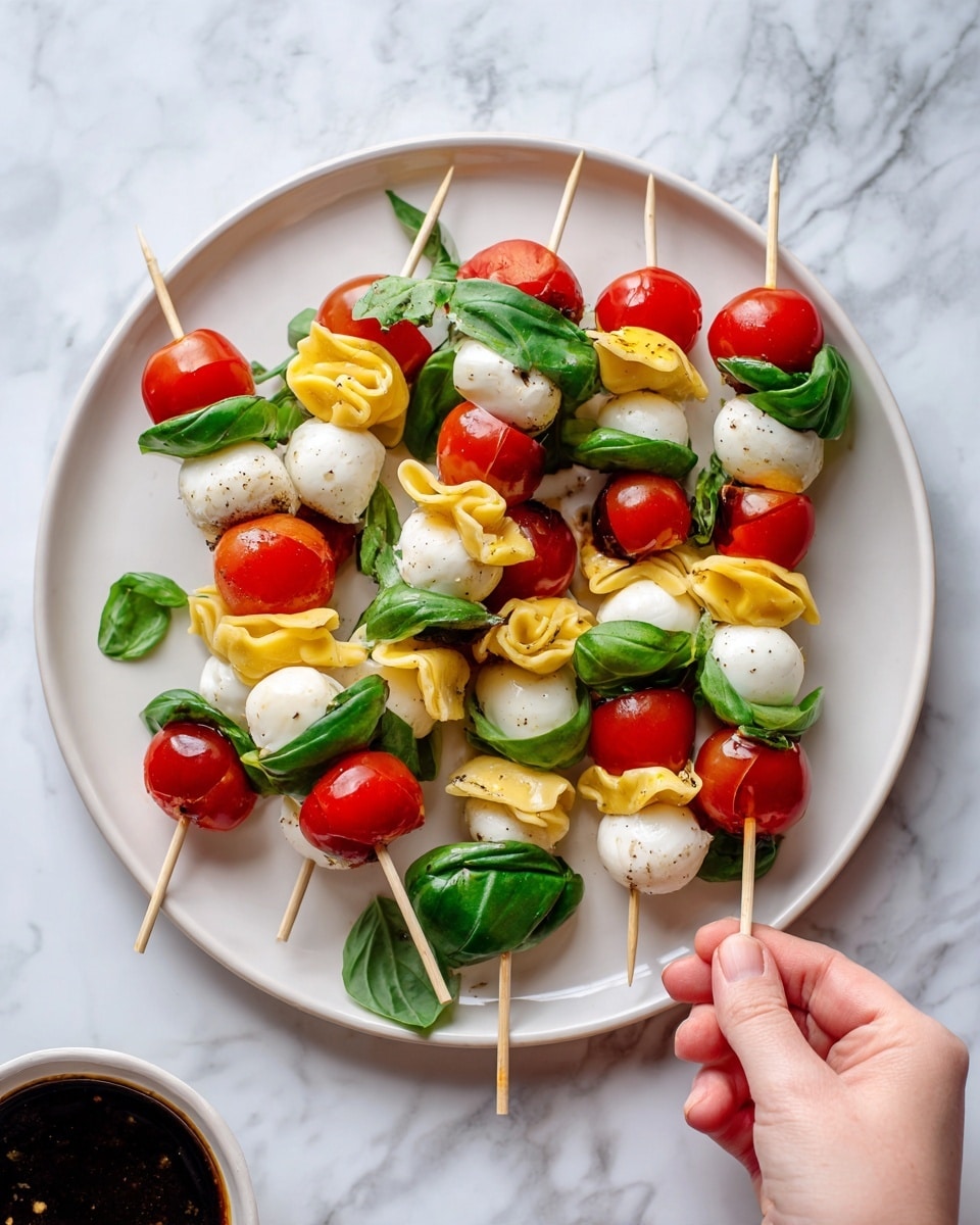 The image shows a white round plate on a white marbled surface, filled with several wooden skewers. Each skewer has three layers: bright green basil leaves on top, followed by red cherry tomatoes in the middle, and white mozzarella balls or folded yellow pasta pieces at the bottom. Some pasta pieces appear stuffed and folded irregularly, adding texture. Extra cherry tomatoes, mozzarella balls, and basil leaves also scatter around the plate and surface. A small white bowl with a dark dip is partially visible at the bottom corner. A woman's hand holds one skewer at the top left of the plate. Photo taken with an iphone --ar 4:5 --v 7