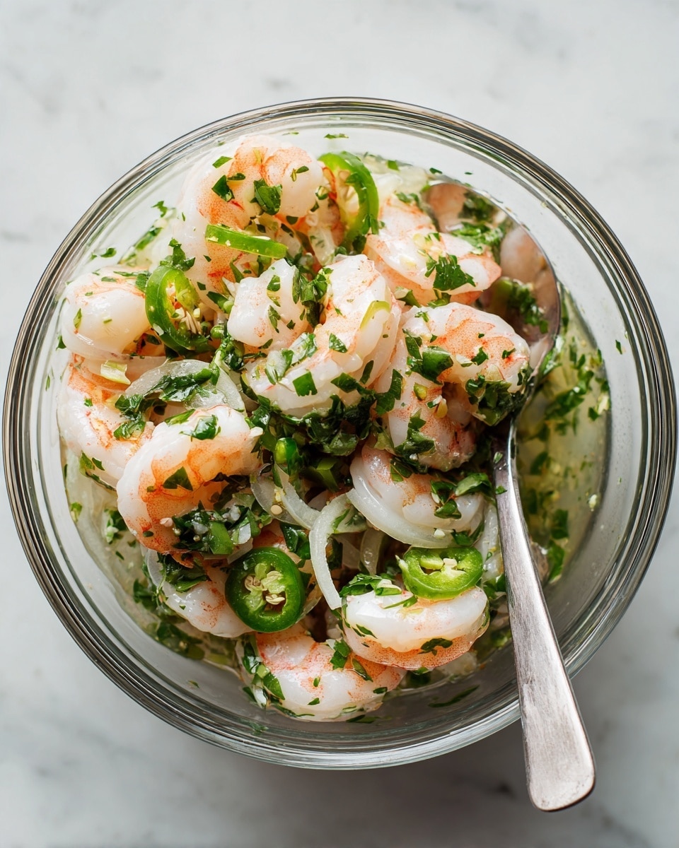 A clear glass bowl holds a vibrant shrimp salad featuring several layers of thick white and pink shrimp. Mixed among the shrimp are thin slices of light green jalapeños and translucent rings of white onion. Sprinkled liberally across the dish are fresh, bright green chopped herbs, likely cilantro, creating a lively contrast. The textures are juicy and fresh, with the shrimp appearing tender and slightly glossy, sitting in a light, clear marinade visible at the bottom of the bowl. A simple metal spoon rests inside the bowl, ready to serve. The bowl is set on a white marbled surface, enhancing the freshness of the salad. photo taken with an iphone --ar 4:5 --v 7