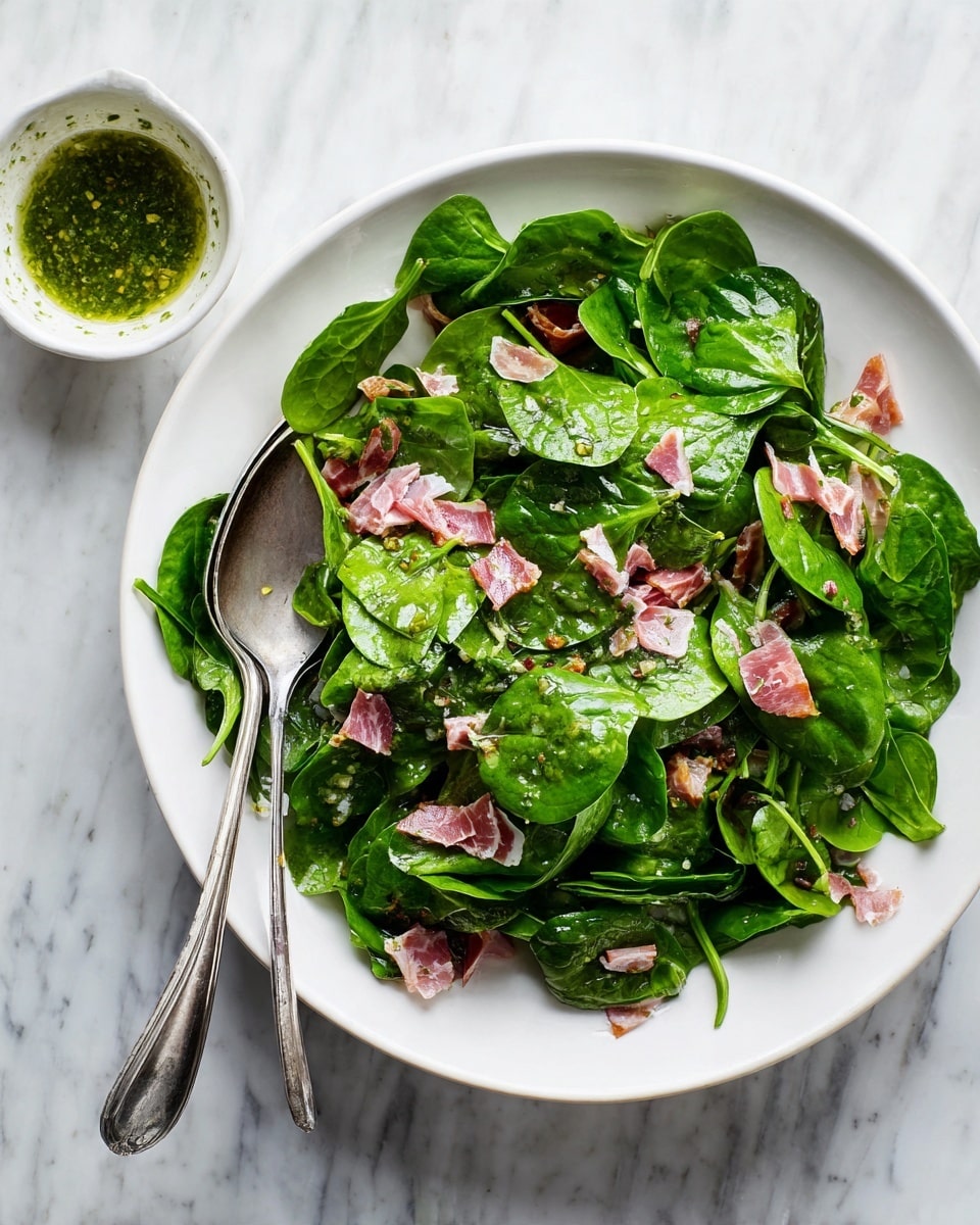 A white round plate filled with a fresh green salad made of many bright spinach leaves spread evenly, mixed with small pieces of thin, pink cured meat scattered throughout. A pair of silver tongs rests on top of the salad. Next to the plate is a small white bowl with green sauce, and a silver spoon inside. All placed on a white marbled surface. photo taken with an iphone --ar 4:5 --v 7