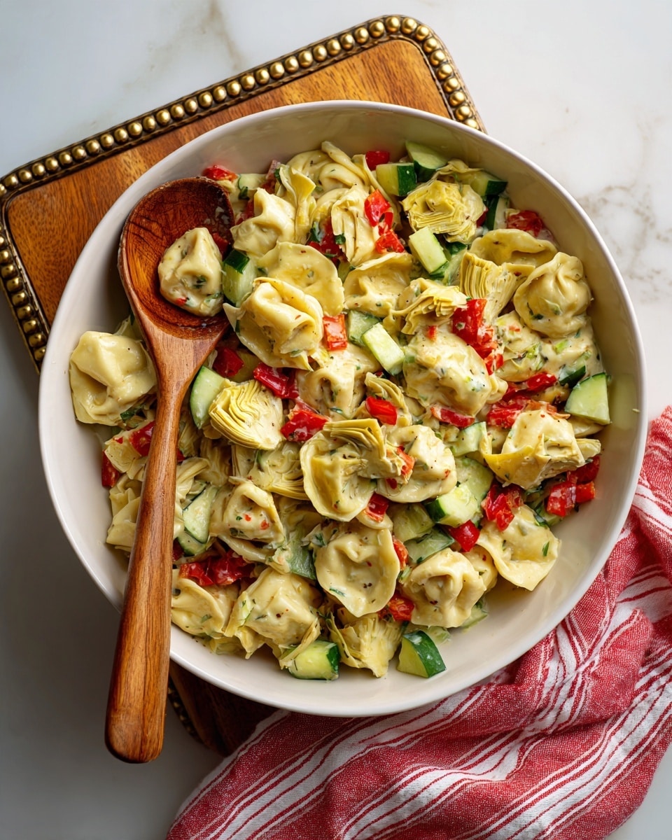 A white bowl filled with a pasta salad sits on a wooden serving board with a gold beaded edge, placed on a white marbled surface. The salad has three main layers: the bottom layer is soft, light beige tortellini pasta; scattered evenly throughout are small pieces of bright green cucumber and diced red bell pepper; on top and mixed in are tender artichoke hearts in pale yellowish-green. A wooden spoon rests inside the bowl, showing the textures and colors of the salad. A red and white striped cloth is casually placed behind the bowl. Photo taken with an iphone --ar 4:5 --v 7